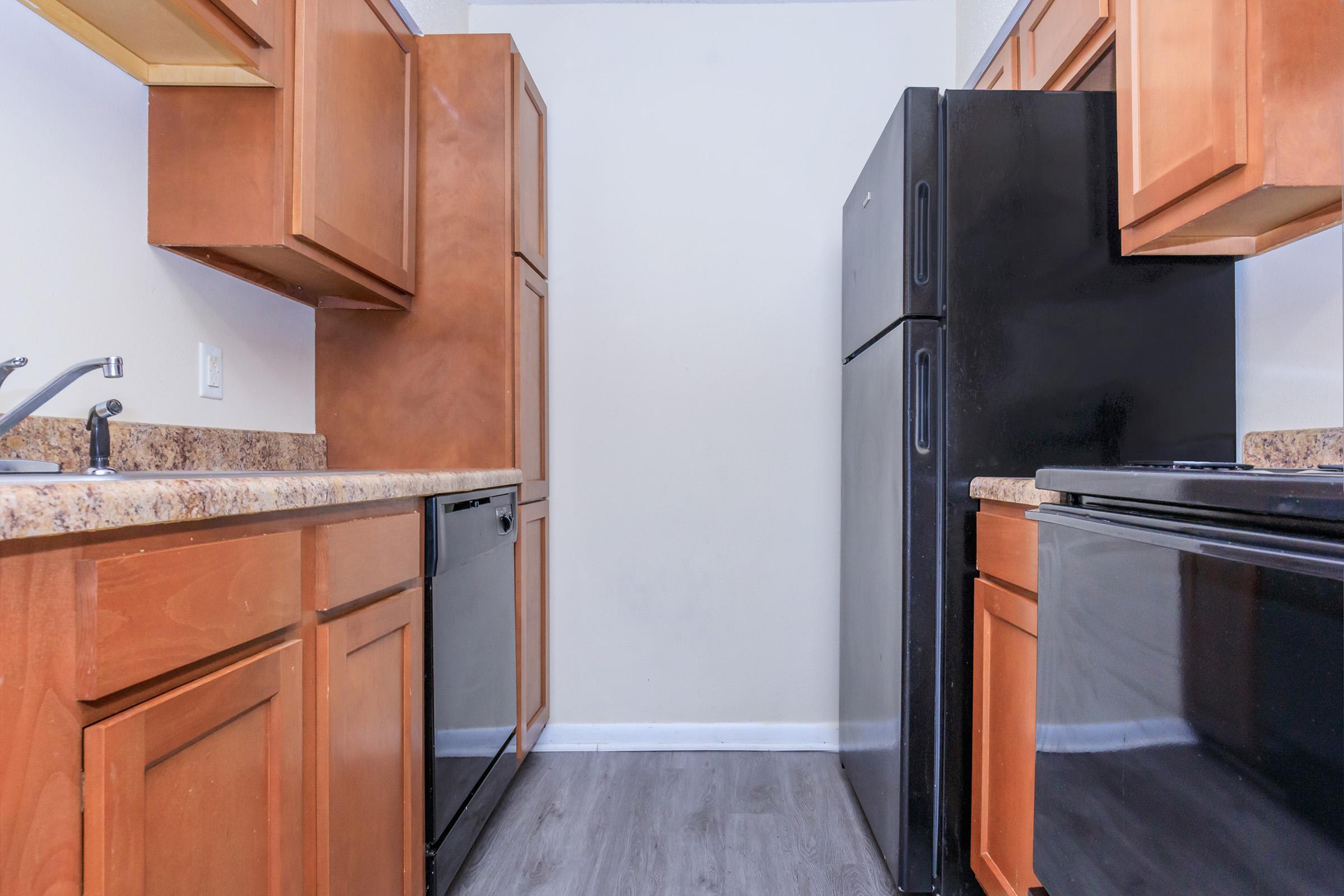 A modern kitchen featuring wooden cabinets, a black refrigerator, and a black oven. The countertop is made of speckled stone, and there is a sink with a faucet on the left. The walls are painted in a light color, and the floor has a dark, textured appearance. The space is well-lit and organized.