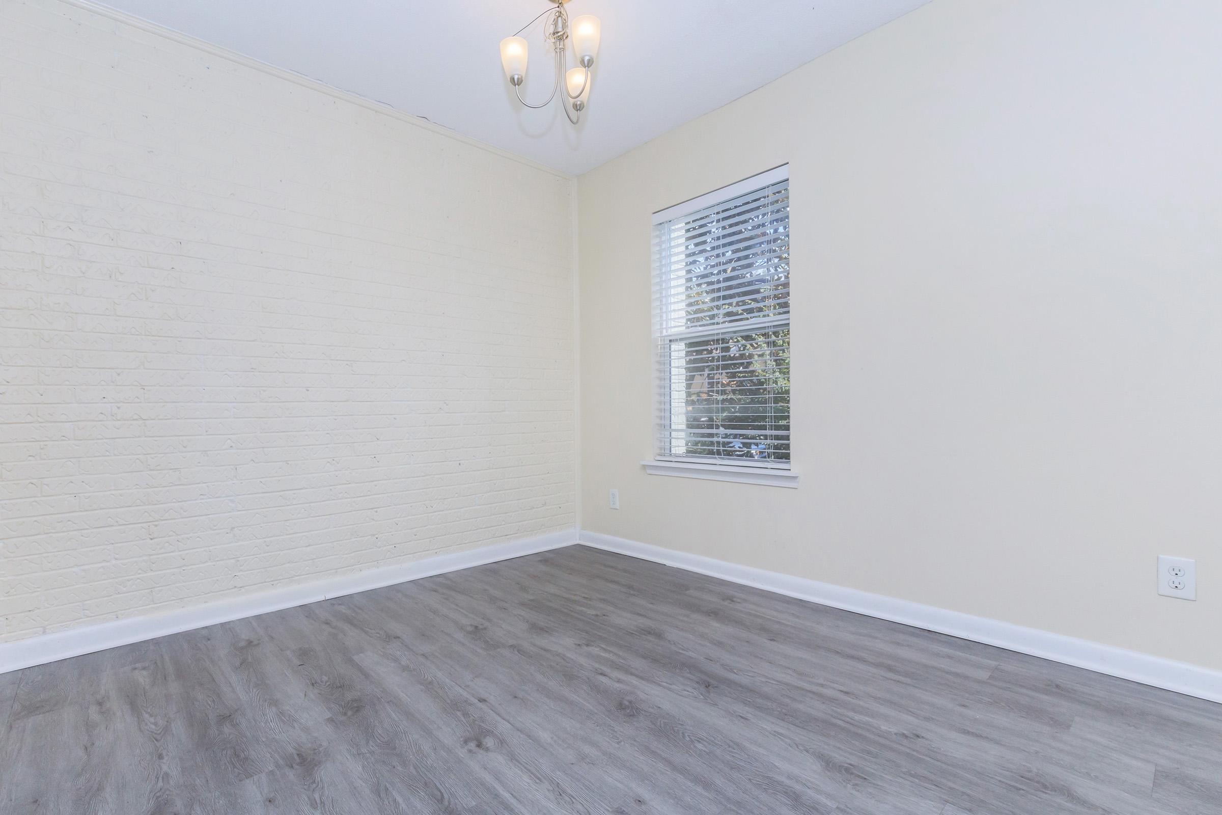An empty room with light-colored walls and a white brick accent wall. There is a window with white blinds allowing natural light, and a modern chandelier hanging from the ceiling. The floor is a light wood laminate, creating a bright and open atmosphere.