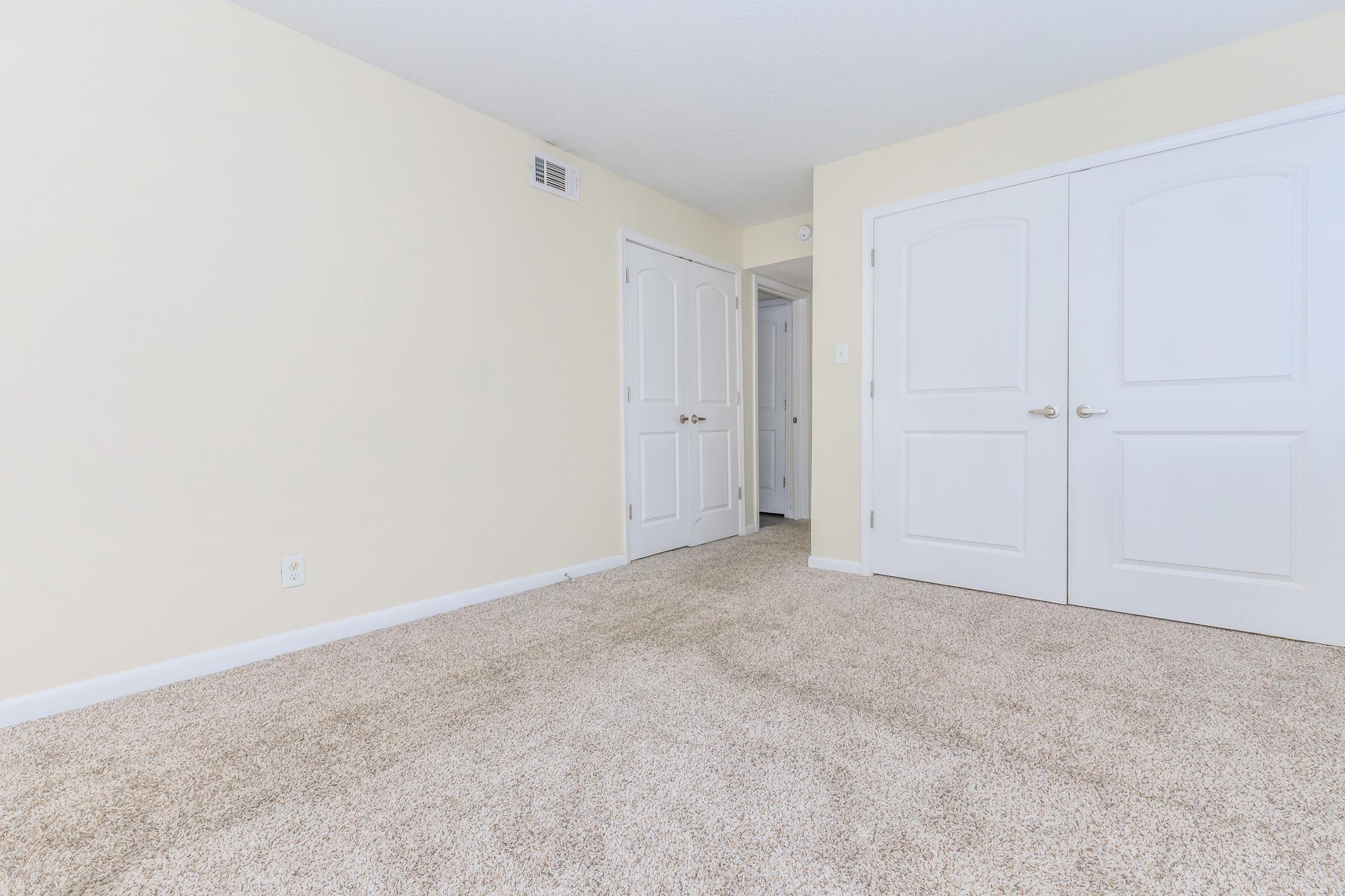 A spacious room with light beige walls and carpeted flooring. Two white double doors are prominent, leading to a closet or adjacent area. The room is well-lit and features a neutral color palette, enhancing its openness and inviting atmosphere.