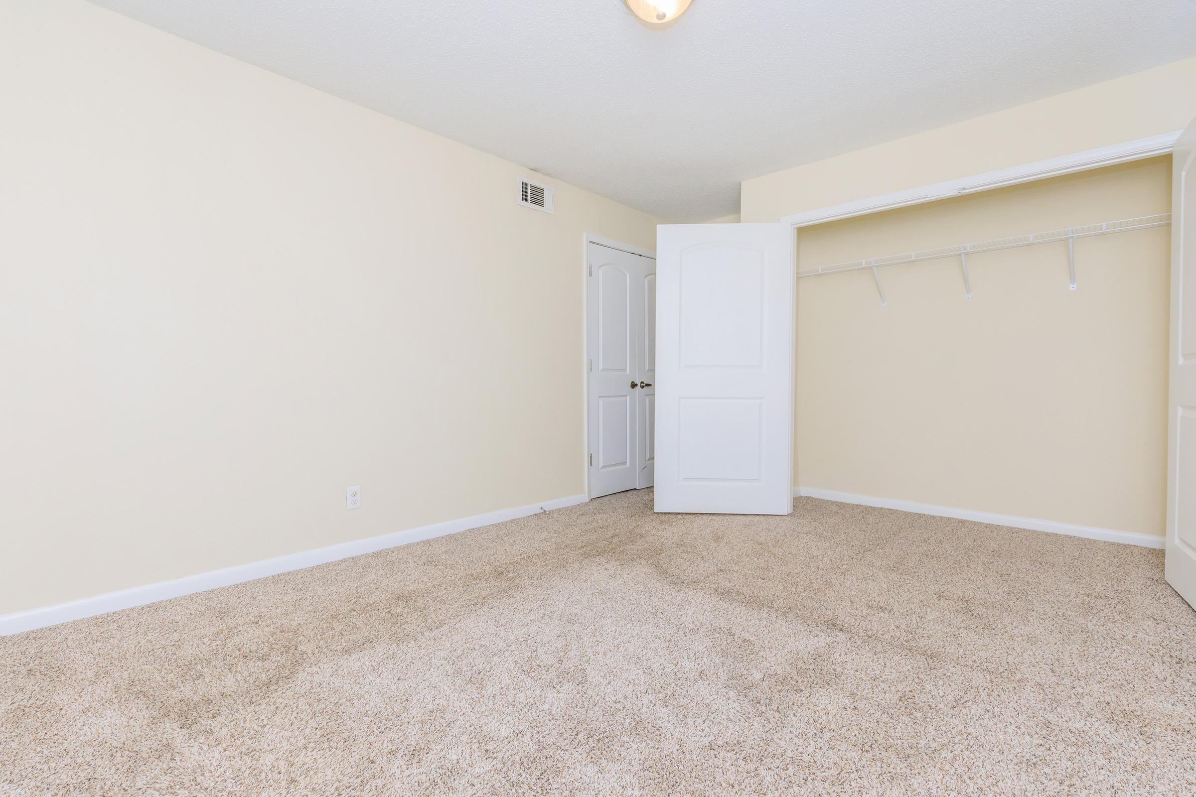 Empty room with beige walls and carpet, featuring a white closet with a single hanging rod and a closed white door. Soft lighting comes from a ceiling fixture. The space appears clean and well-maintained, suitable for various purposes.