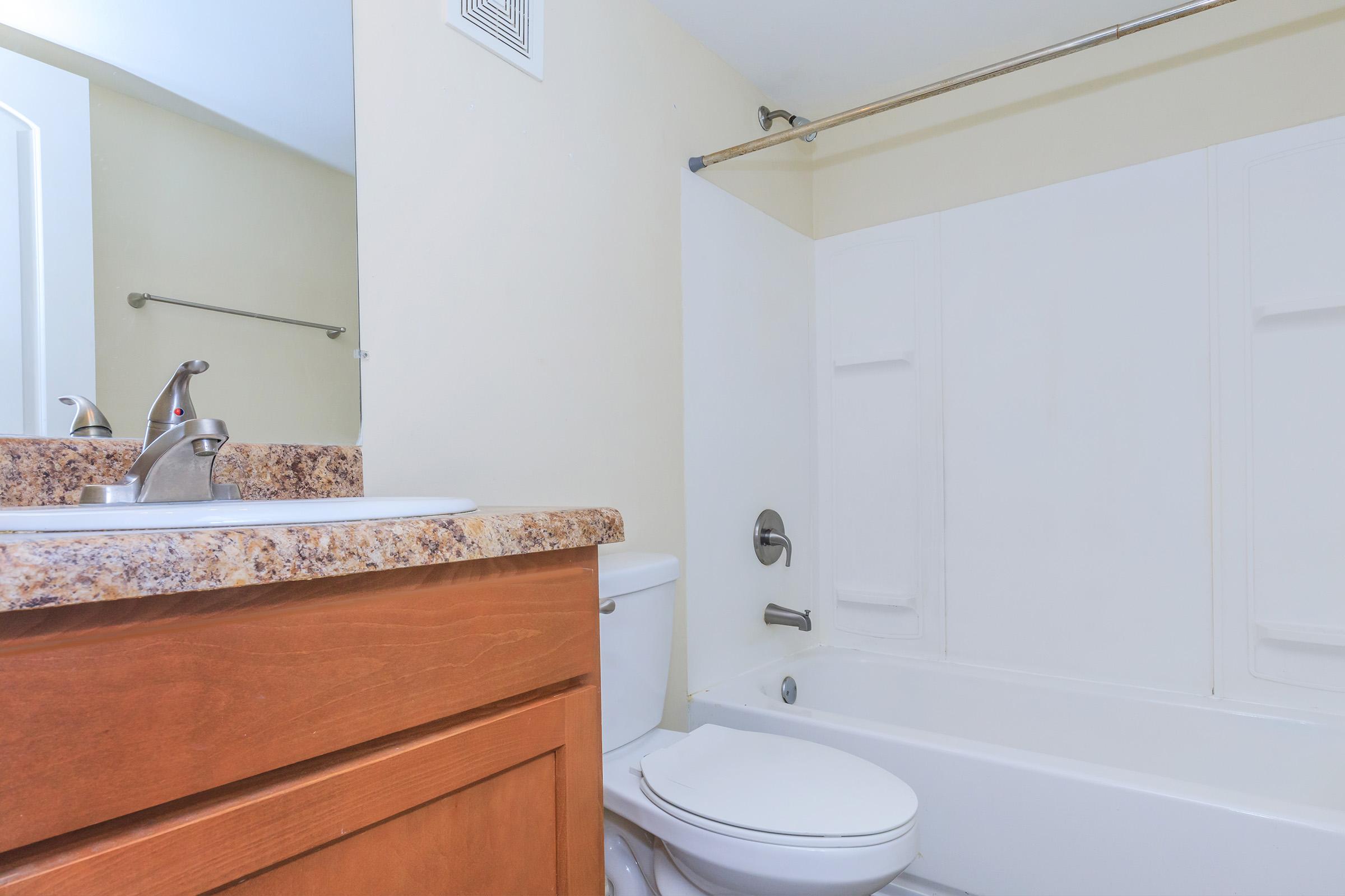 A clean bathroom featuring a bathtub with a shower curtain, a toilet, and a sink with a granite countertop. The walls are painted a light color, enhancing the spacious feel. A mirror above the sink reflects the room, and a towel bar is mounted on the wall.