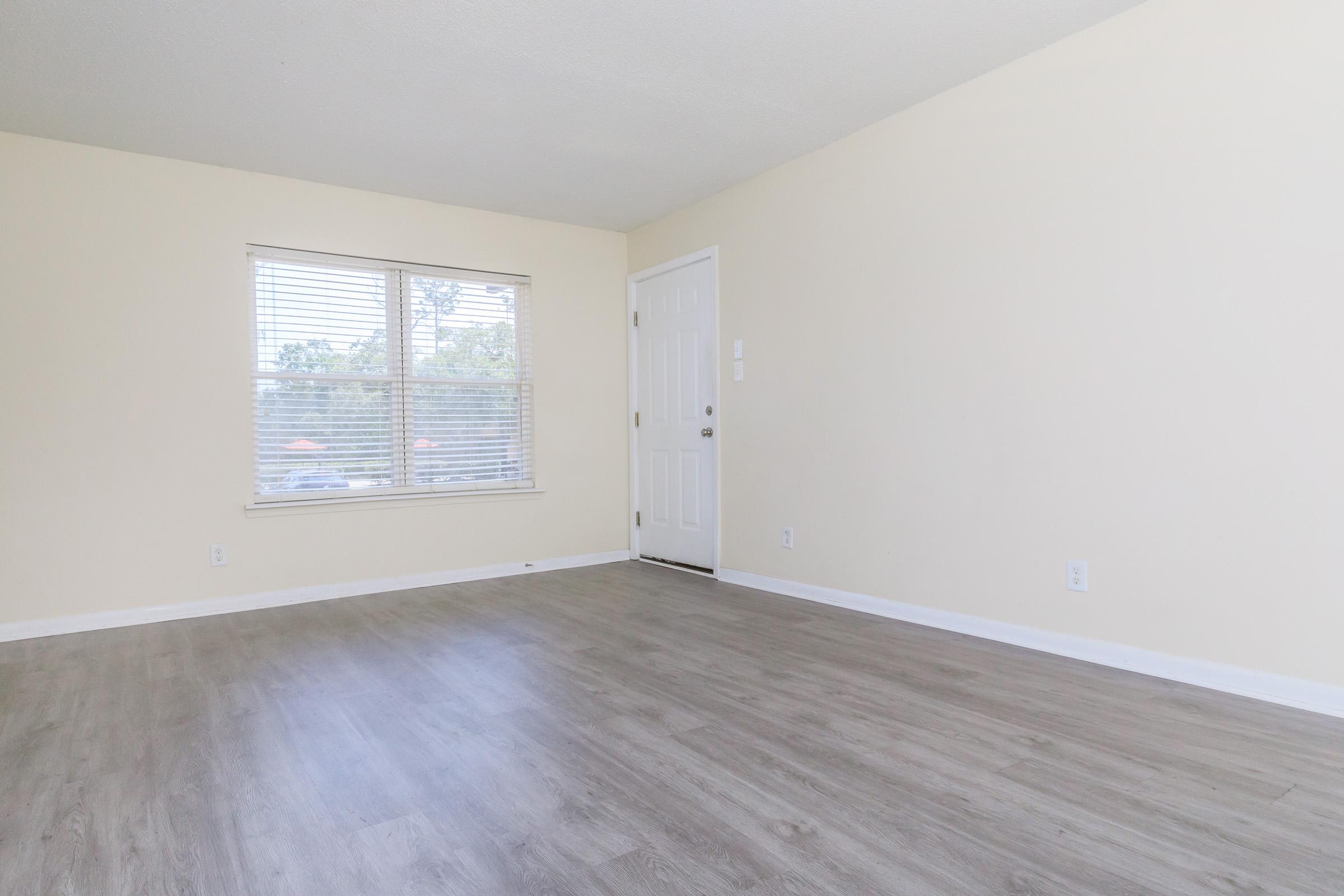 A bright, empty interior room with light-colored walls and a smooth wooden floor. It features a window with horizontal blinds allowing natural light and a white door leading outside. The space is minimalistic and clean, suitable for various uses or decoration.