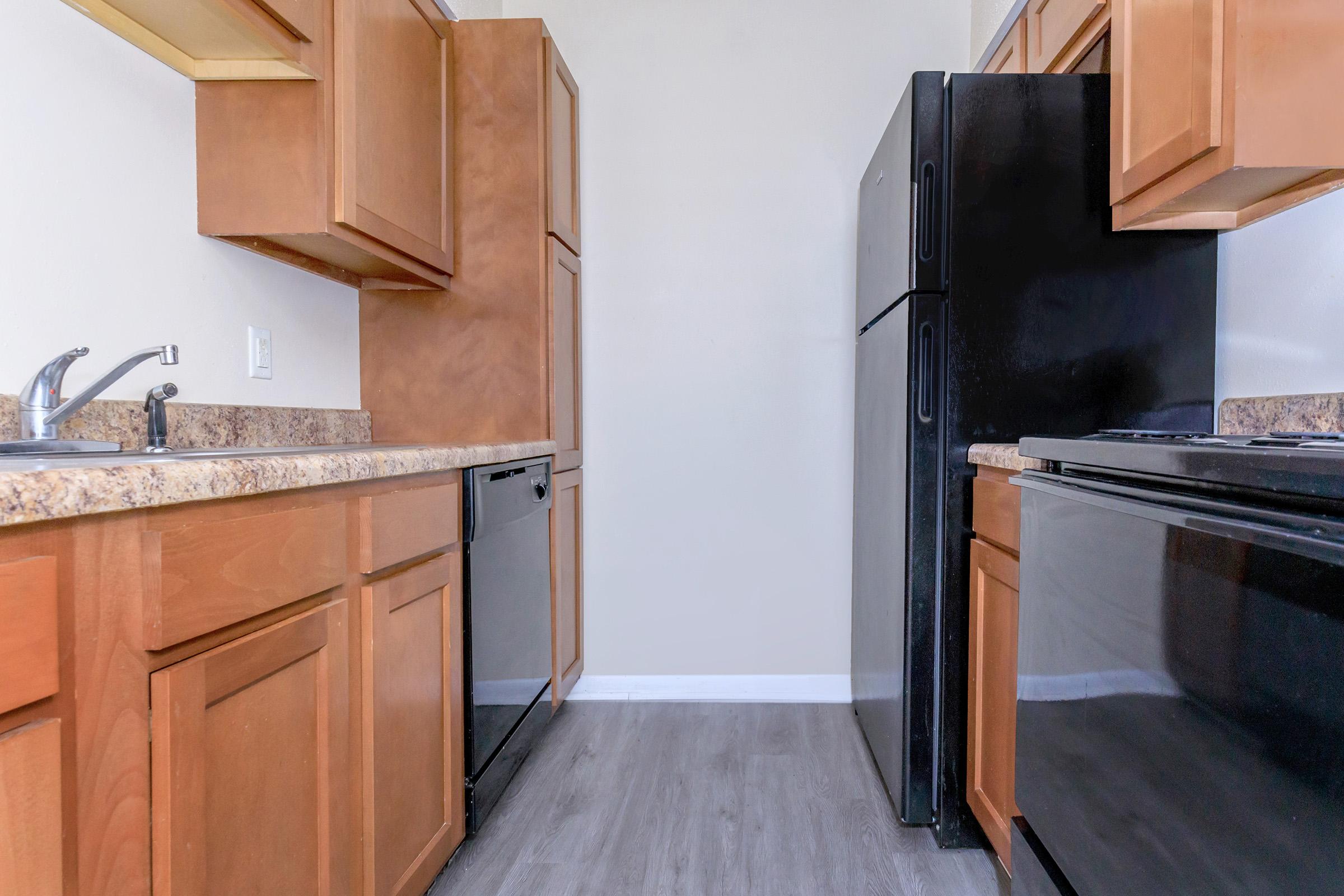 A modern kitchen featuring wooden cabinets, a black refrigerator, a black stove, and a black dishwasher. The countertop is a light-colored stone, and the flooring is a gray wood-like finish. The walls are painted in a neutral shade, creating a bright and spacious atmosphere.