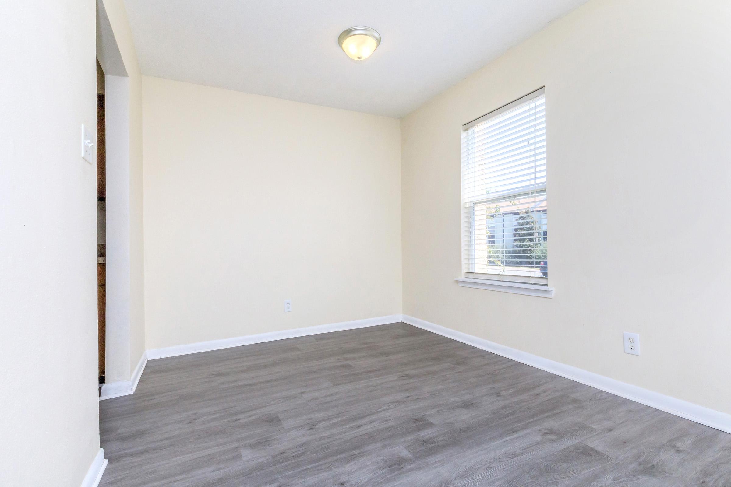 A vacant room with light-colored walls, a window with blinds, and a ceiling light fixture. The floor features light wood-like laminate. There's a doorway leading to another area, creating a spacious and bright atmosphere. Ideal for a living space or office.