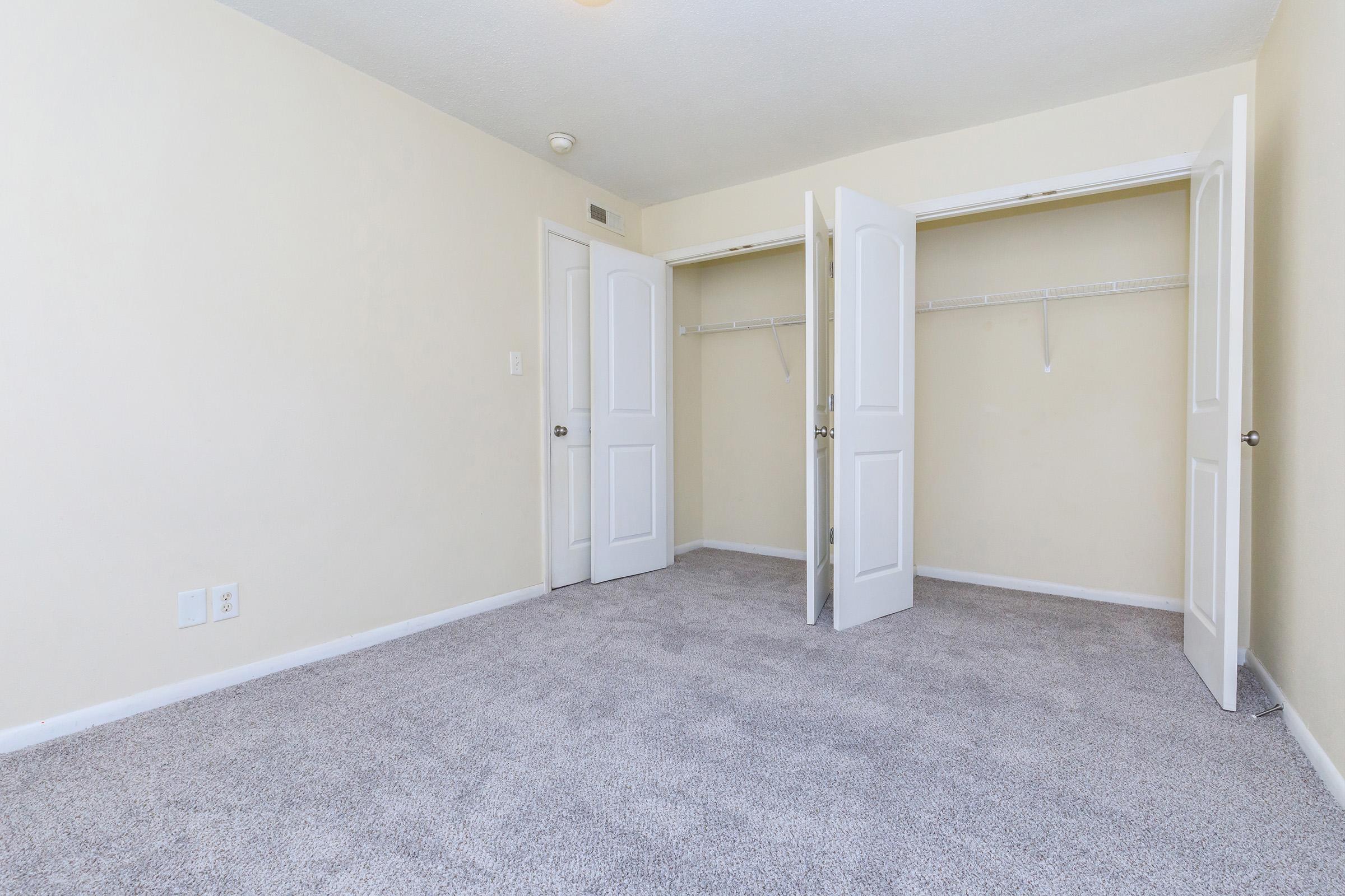 A spacious, empty room with beige walls and light gray carpet. There are two open white doors leading to a closet with hanging space. The room has natural light and no furniture, making it appear bright and inviting.