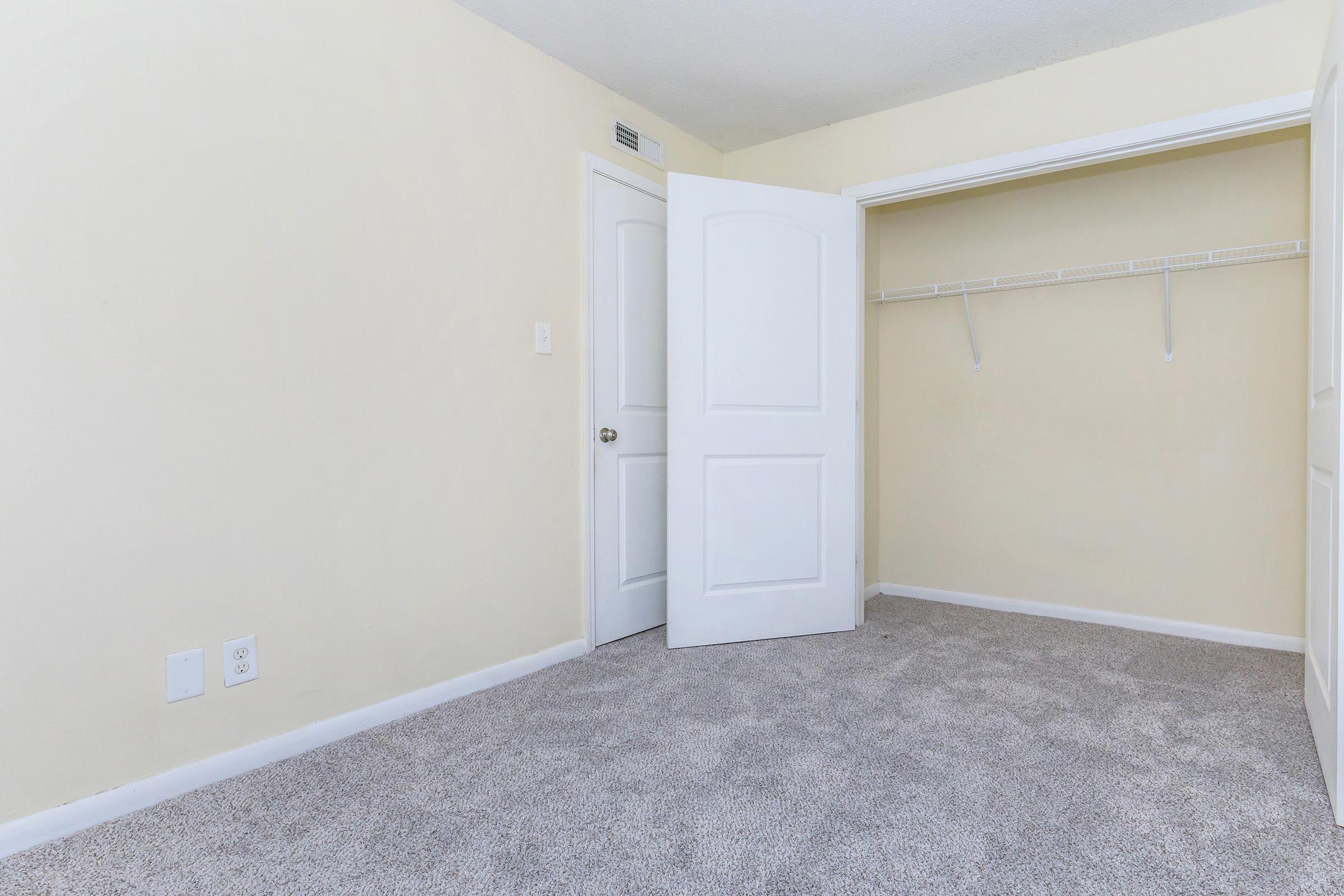 A vacant room with light yellow walls and beige carpet. One white door is slightly ajar, leading to another area. The closet on the right features a hanging rod and no shelves. The space is well-lit, giving a clean and open feel.