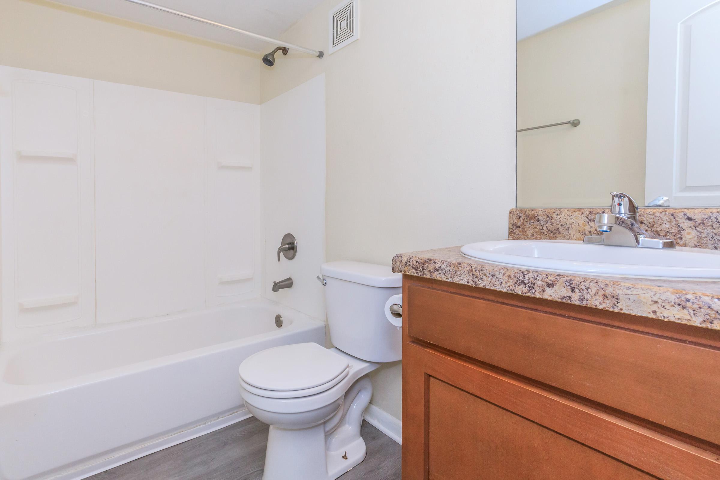 A clean and simple bathroom featuring a bathtub with a shower, a toilet, and a sink with a granite countertop. Light-colored walls and a large mirror above the sink create a bright atmosphere. The flooring is a modern, light grey.
