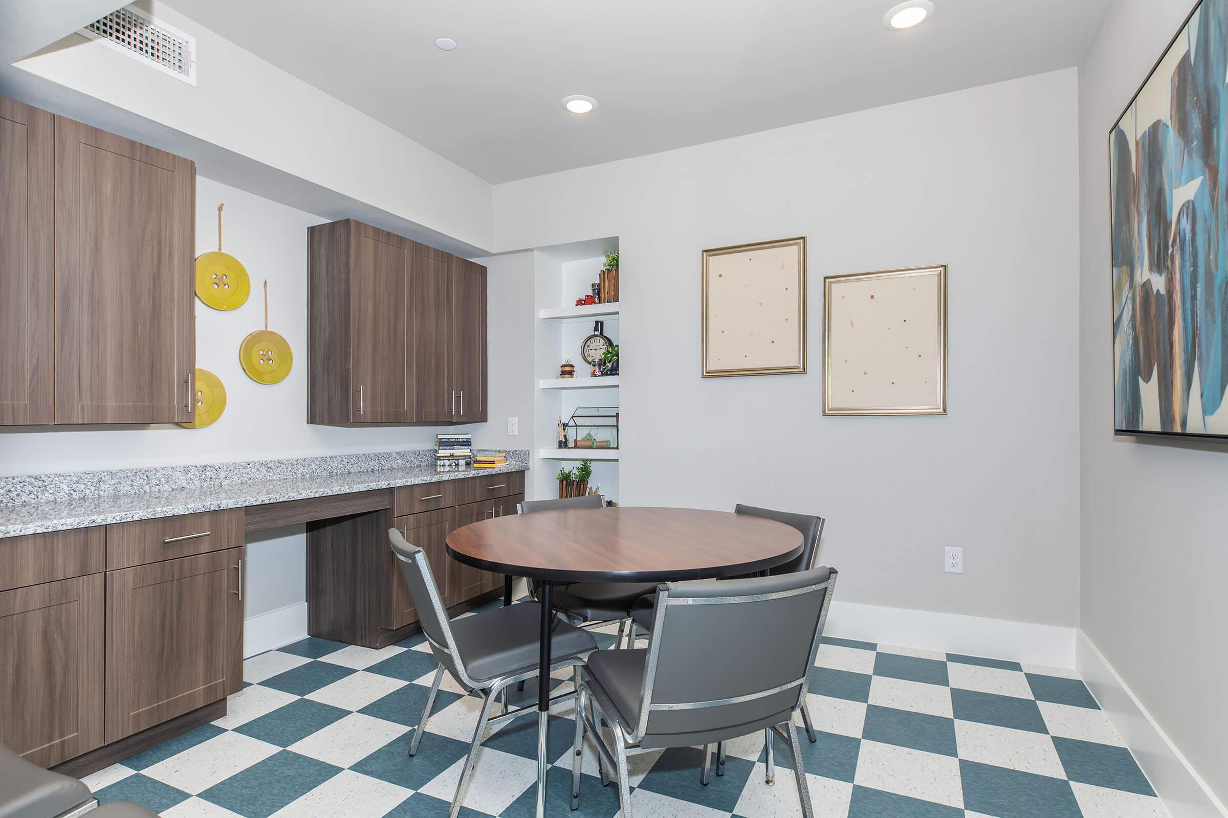 A modern kitchen or dining area featuring a round wooden table with chairs, stylish cabinetry, decorative shelves, and wall art. The floor is covered with black and white checkered tiles, and the space is well-lit with recessed lighting.