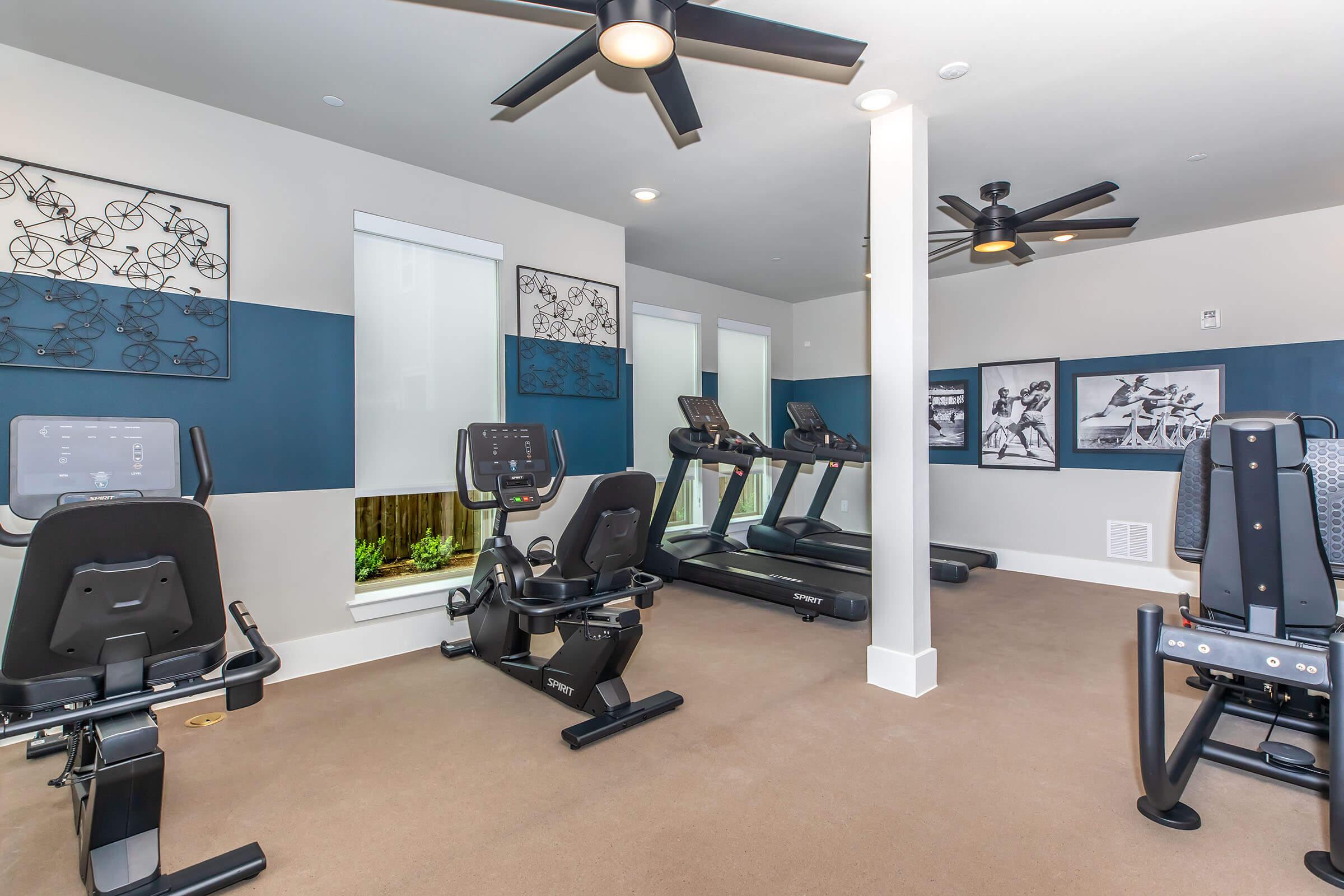 A modern gym or fitness room featuring several exercise machines, including treadmills and stationary bikes, with large windows allowing natural light. The walls are decorated with artwork, and the space has a clean, organized layout with ceiling fans for ventilation.