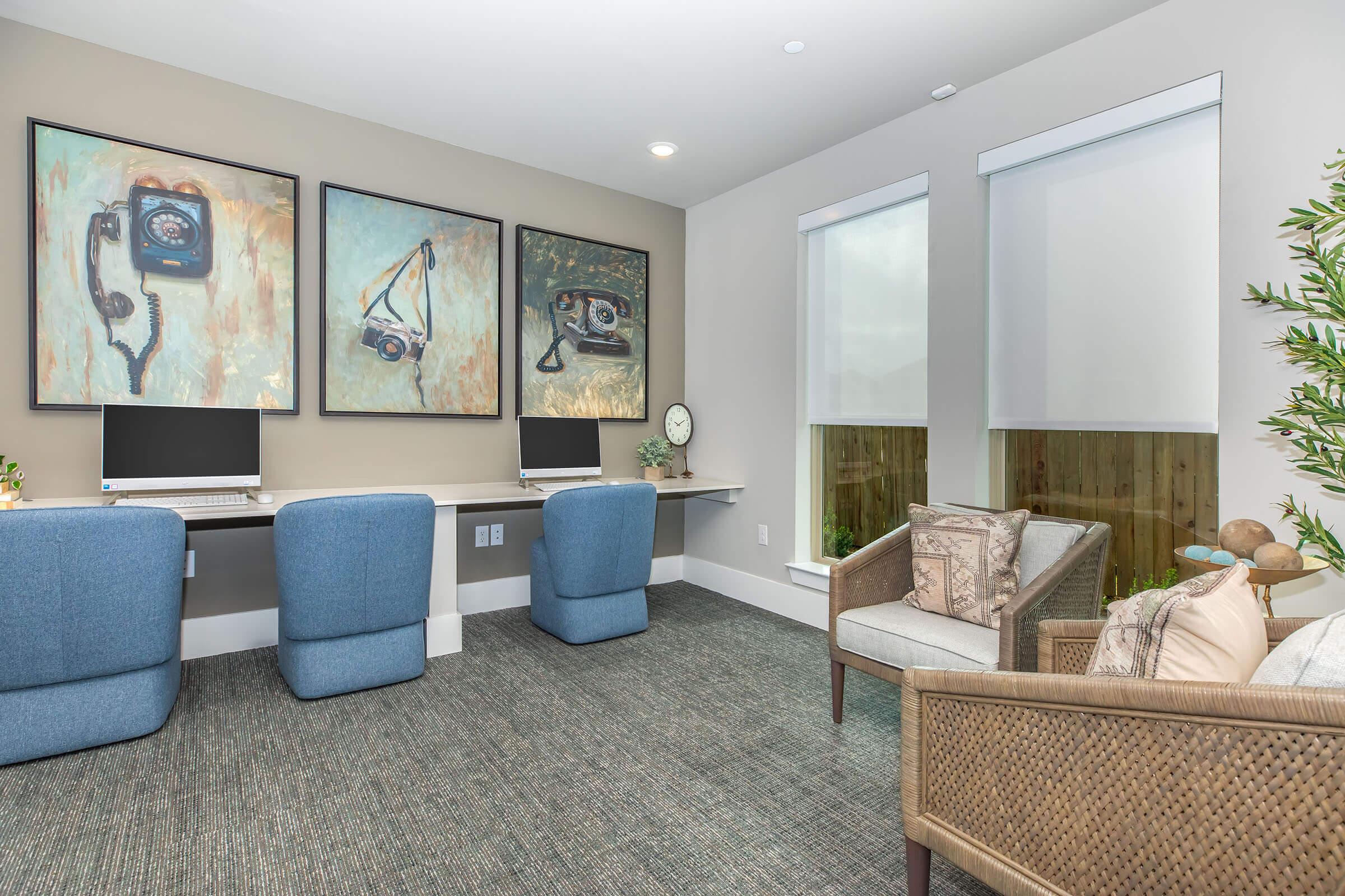 A modern home office featuring two desks with computers, framed artwork of vintage cameras on the wall, and a cozy seating area with a wicker chair and cushions. Natural light filters through window shades, creating a bright and inviting workspace. Decor includes a plant and a decorative clock.