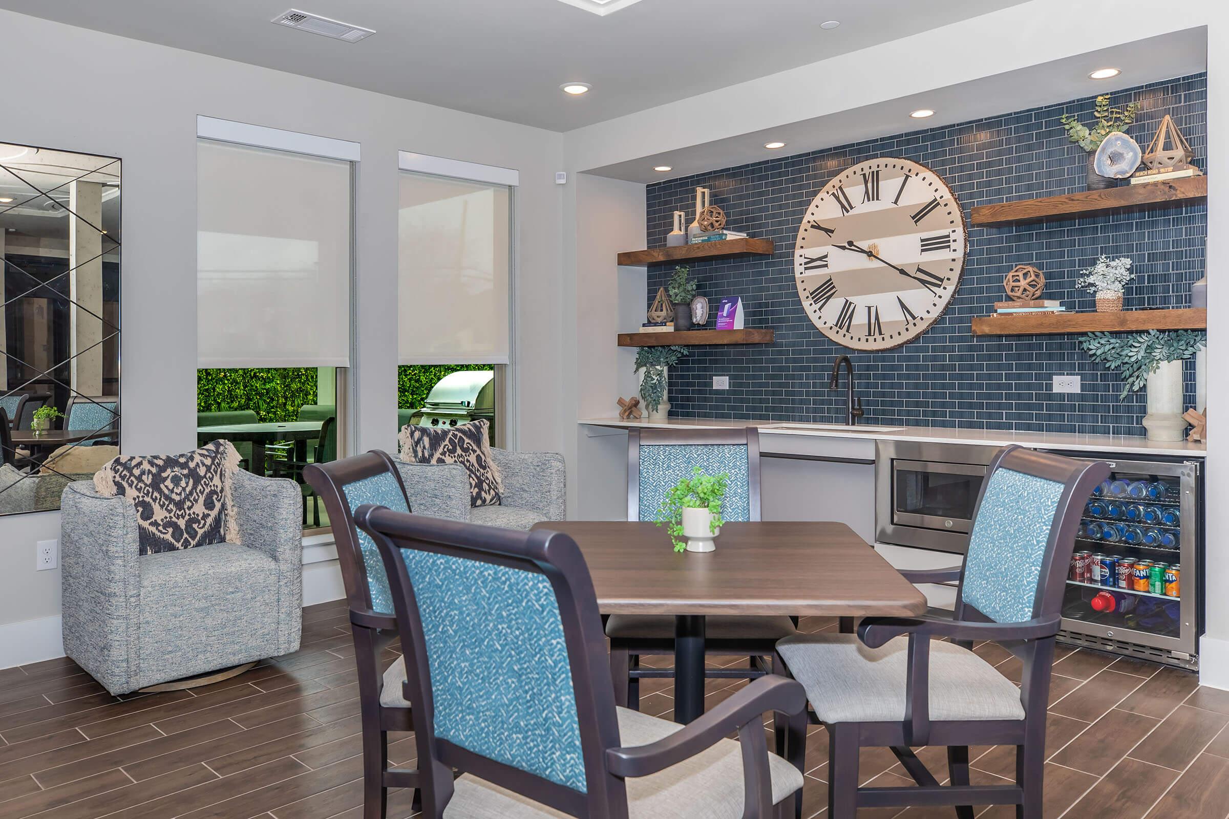 A modern dining area featuring a circular wall clock, wooden shelves displaying decorative items, and a kitchenette. The table is surrounded by four chairs with blue upholstery. Large windows allow natural light, and a green outdoor view is visible through the glass. The decor is contemporary and inviting.
