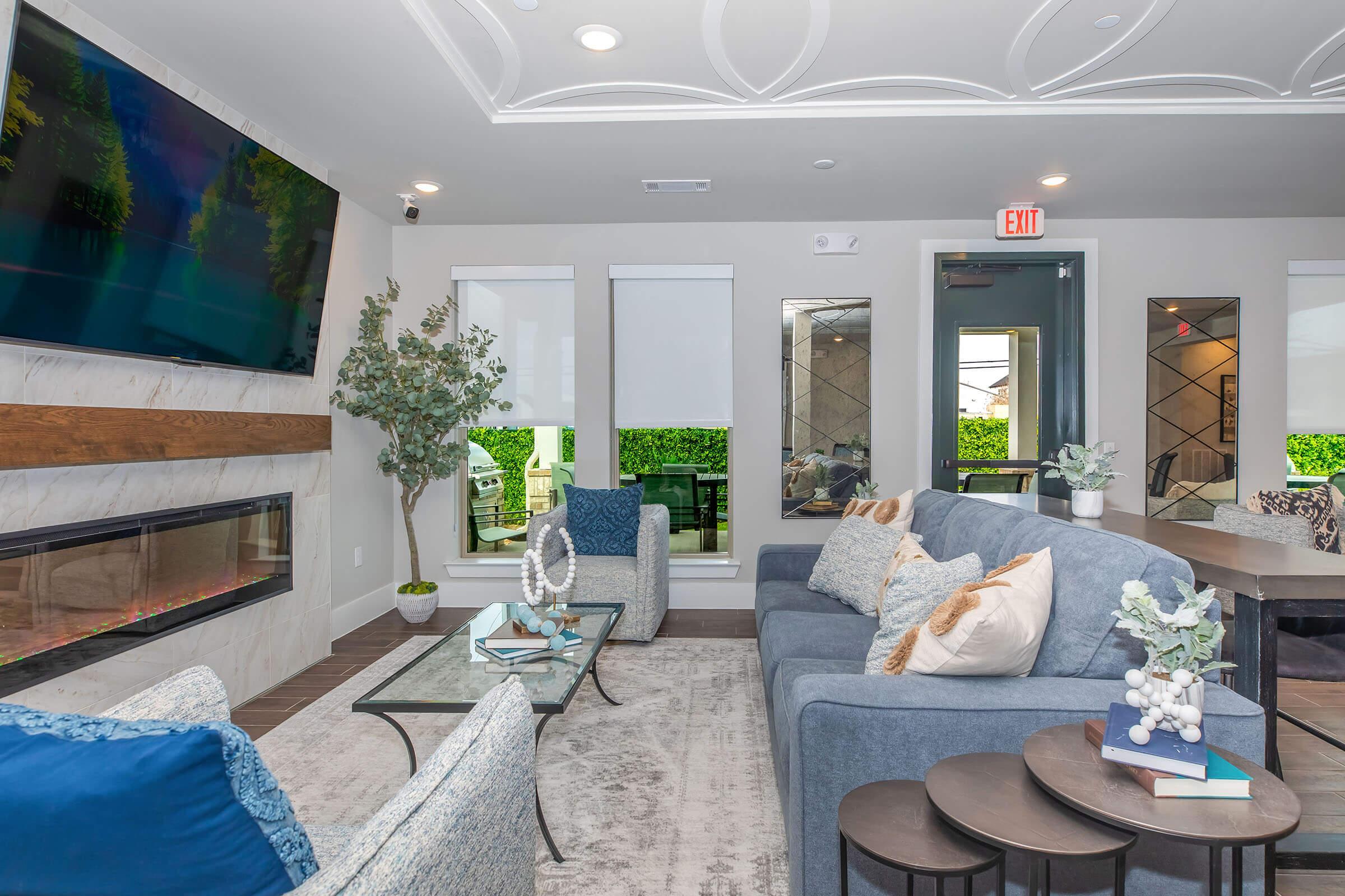 A modern living room featuring a blue sofa with decorative pillows, a glass coffee table, and a sleek fireplace. There’s a large TV on the wall, decorative plants, and large windows providing a view of greenery. The space has a stylish, contemporary design with soft lighting.