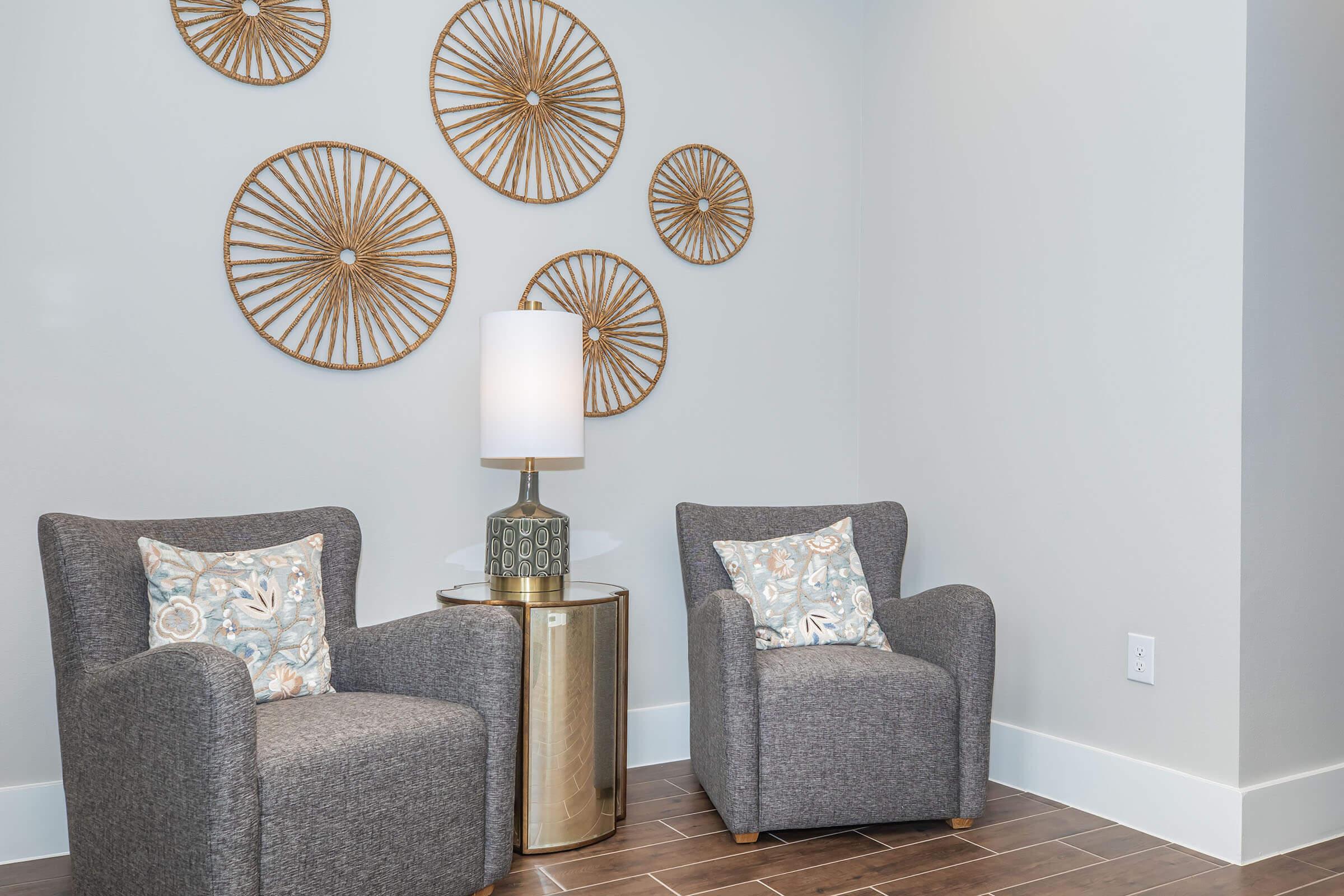 Two cozy gray armchairs with decorative pillows are positioned near a golden side table and a modern lamp. The wall features circular, woven wall art, creating a stylish and inviting atmosphere. The setting has a light and airy vibe, complemented by wooden flooring and soft wall colors.