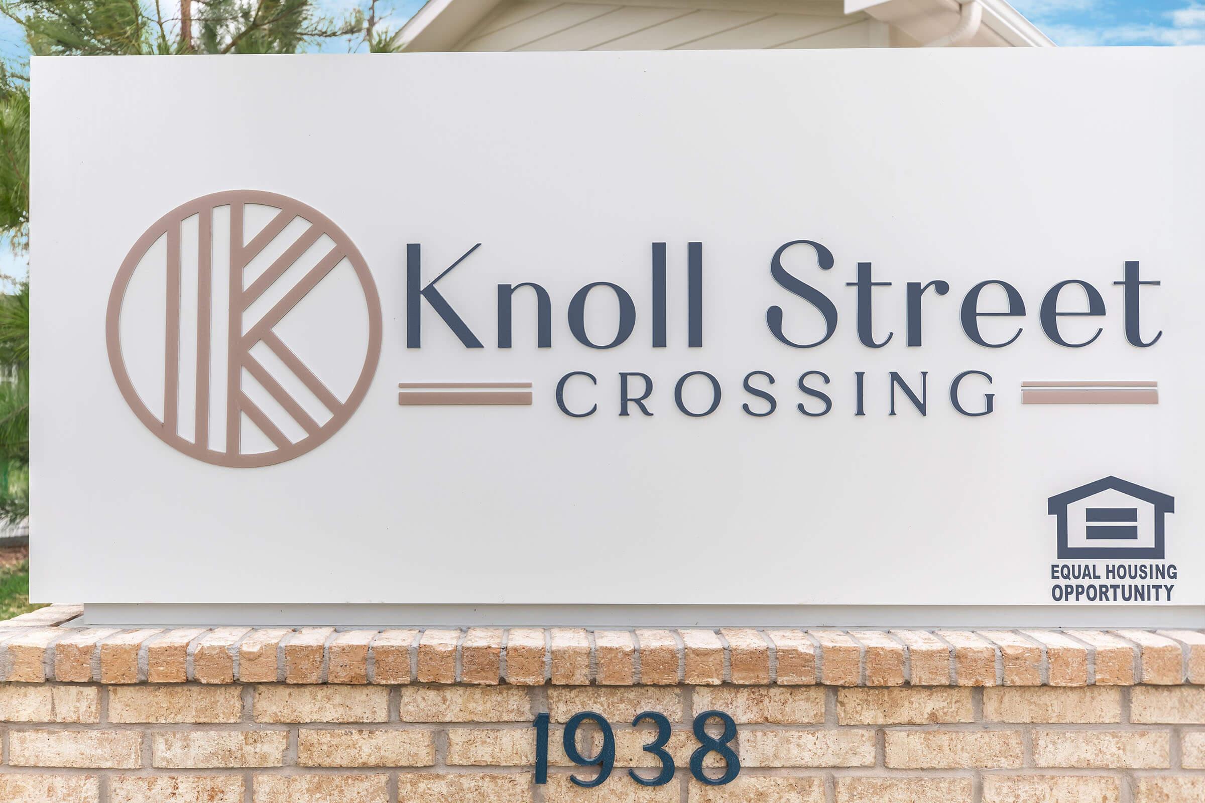 Sign for "Knoll Street Crossing" prominently featuring the name and a logo, with the address "1938" below it. Includes an emblem indicating "Equal Housing Opportunity." The sign is mounted on a brick structure, set against a backdrop of a clear sky and greenery.