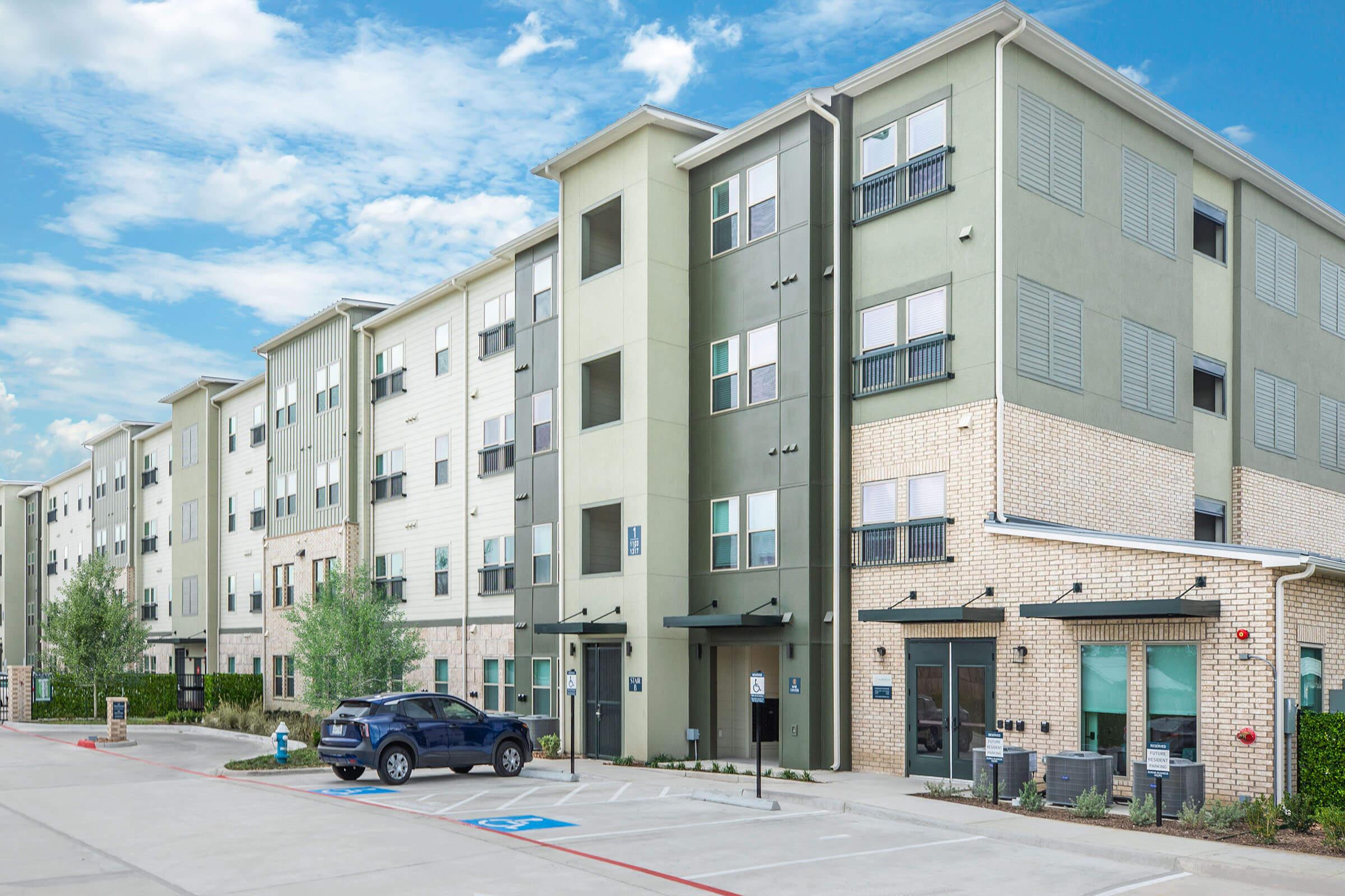 A modern apartment building featuring multiple stories with a mix of light and dark green exteriors and brick accents. The structure has balconies, large windows, and a welcoming entrance. There is a parking lot in front with a vehicle parked, and trees are visible nearby under a blue sky.