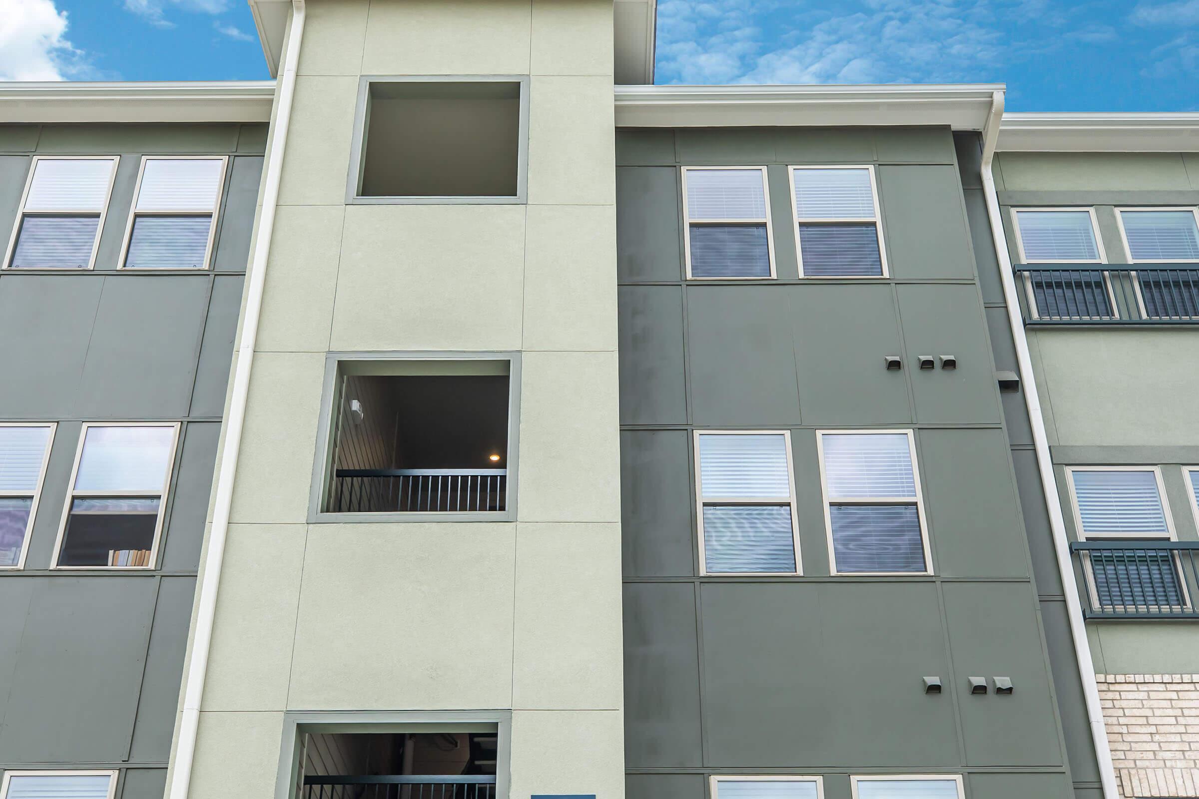 A modern multi-story building featuring a mix of gray and light green exteriors. The structure has several windows, some with black railings, and an empty balcony space. The sky above is partly cloudy, contributing to a contemporary urban aesthetic.