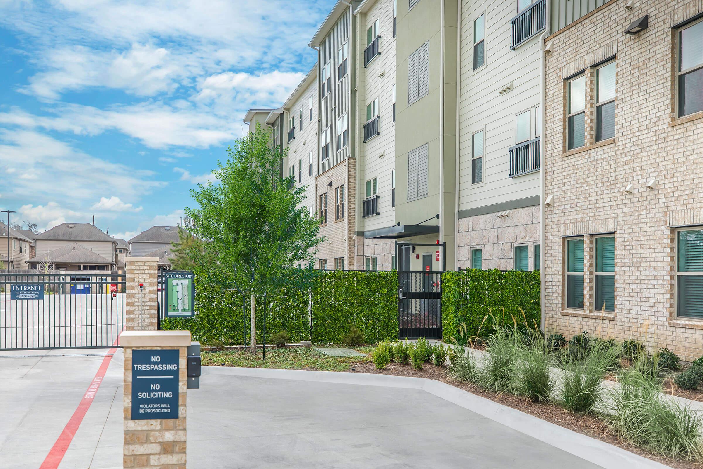 A modern apartment complex with brick and green siding, featuring multiple stories and large windows. In the foreground, there's a well-maintained courtyard with landscaping, a "No Trespassing" sign, and a gated entrance visible in the background. The sky is clear with a few clouds.