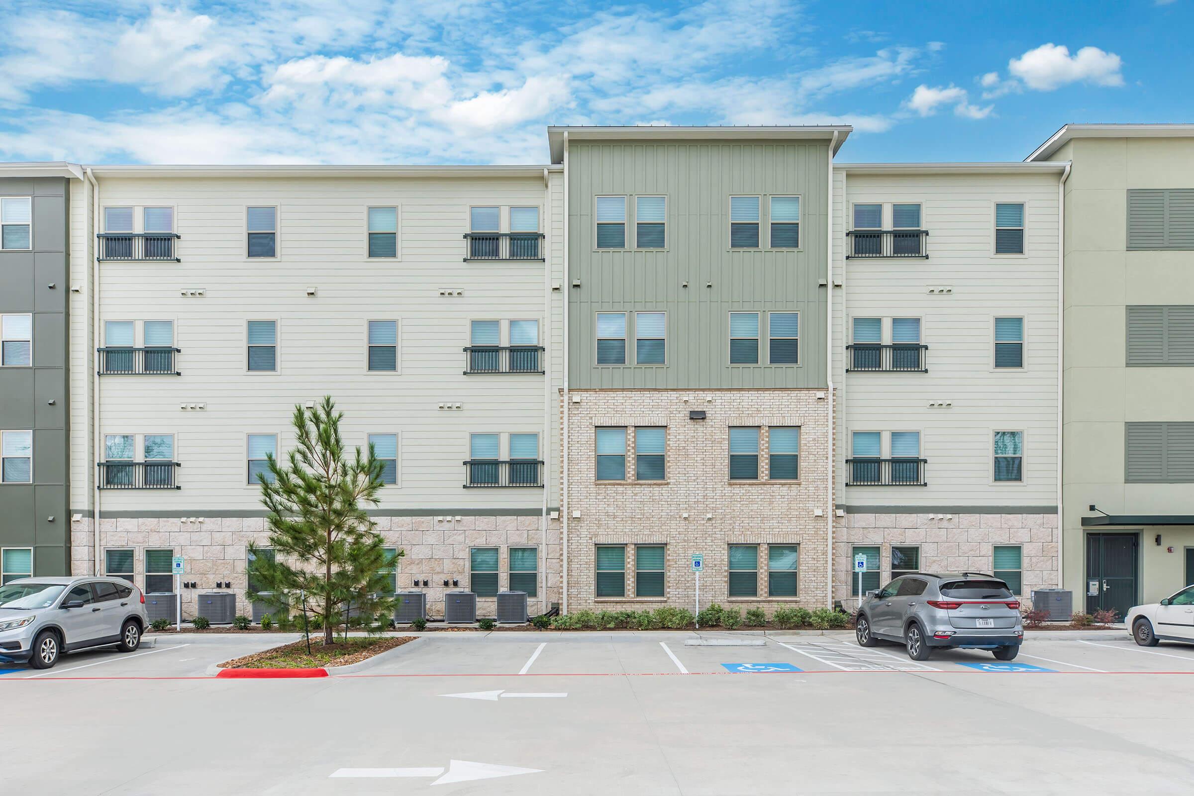 A multi-story apartment building featuring a combination of brick and siding. The façade includes numerous windows, a mix of colors, and air conditioning units. In front, there is a parking lot with several parked vehicles and landscaping elements, including small trees. The sky is clear with a few clouds.