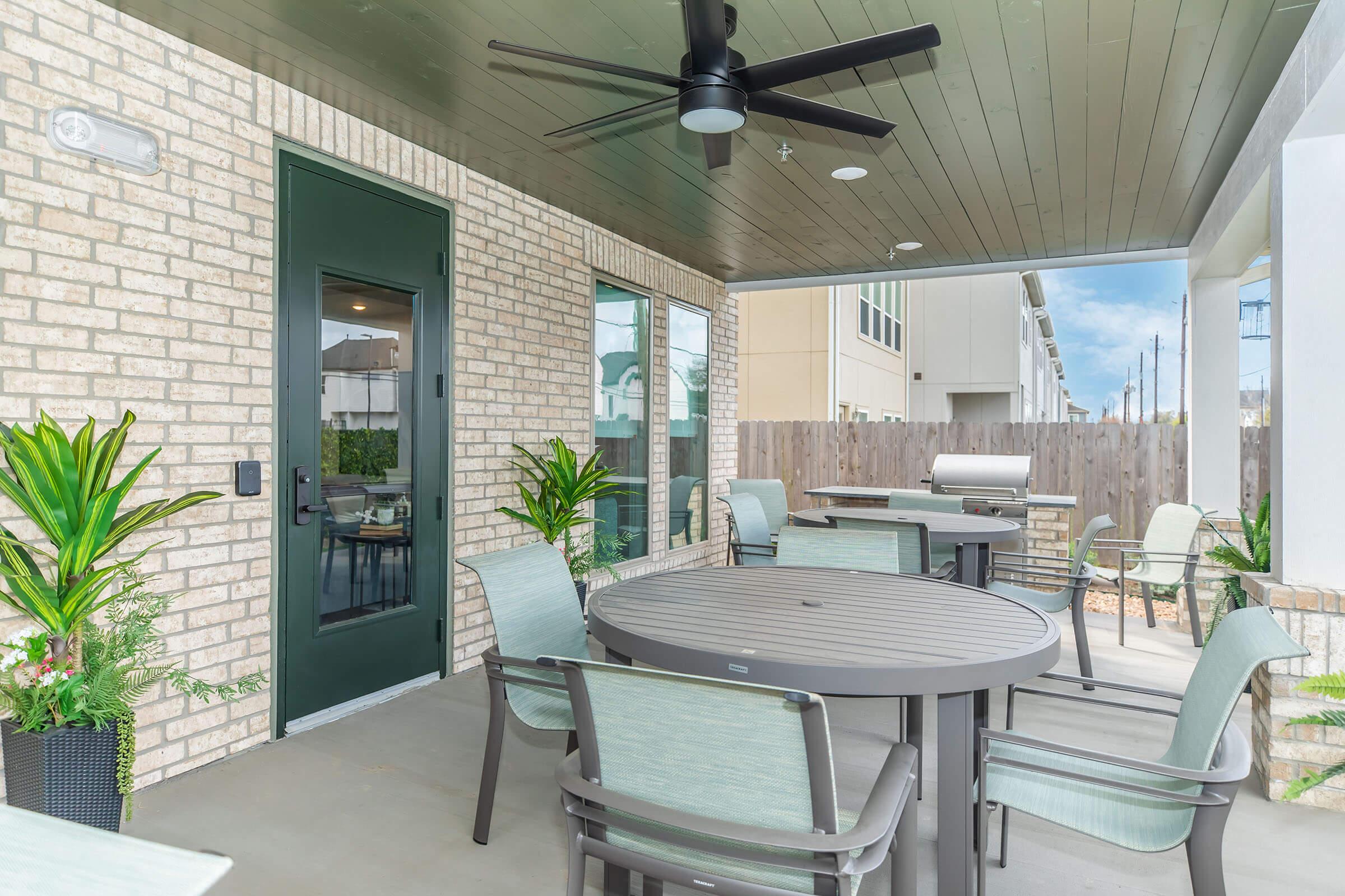 A shaded outdoor patio featuring a round table surrounded by chairs, potted plants for decoration, a ceiling fan, and a grill nearby. The space has brick walls and large windows, creating a welcoming atmosphere for gatherings or relaxation.