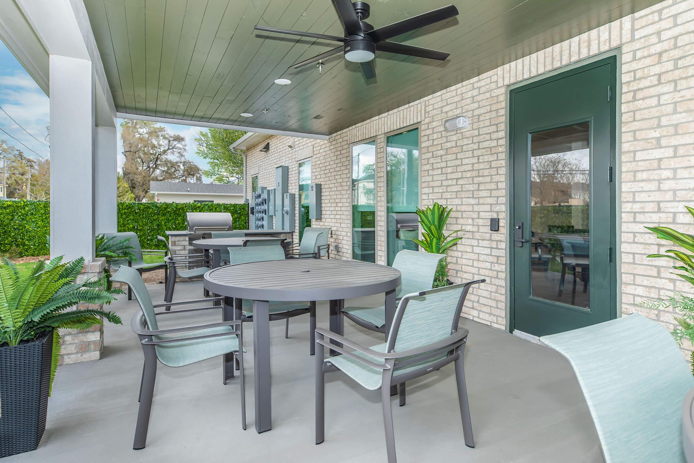 A covered outdoor patio area featuring a round table with four chairs, a ceiling fan, and potted plants. The space is surrounded by brick walls and large glass doors, providing a cozy spot for relaxation or dining. Greenery is visible in the background.