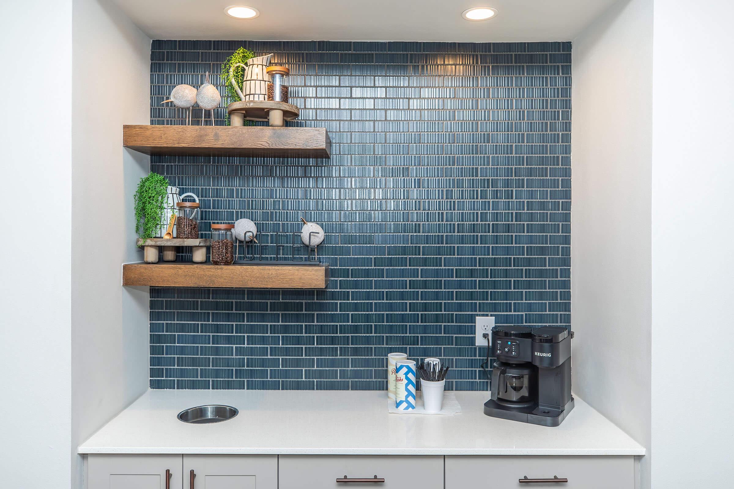 A modern kitchen setup featuring a coffee maker on a white countertop with a circular sink. Above, there are two wooden shelves displaying decorative items and plants against a blue tiled wall. The overall design is clean and contemporary, with integrated lighting.