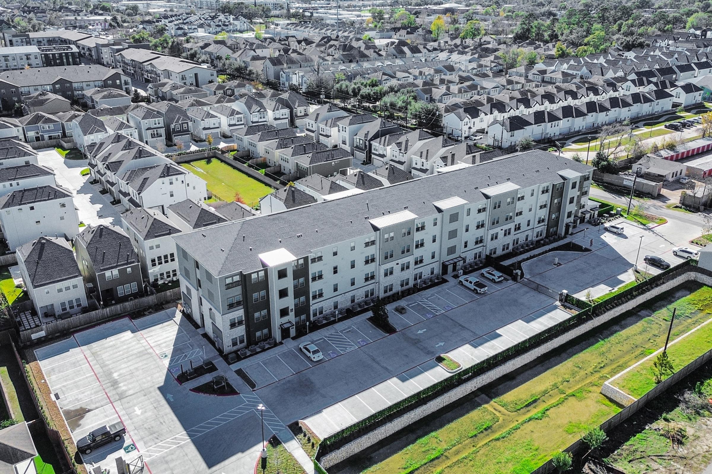 Aerial view of a modern apartment complex surrounded by residential neighborhoods. The complex features multiple stories with a parking lot in front, while the background includes rows of homes and greenery, showcasing urban development and suburban living.