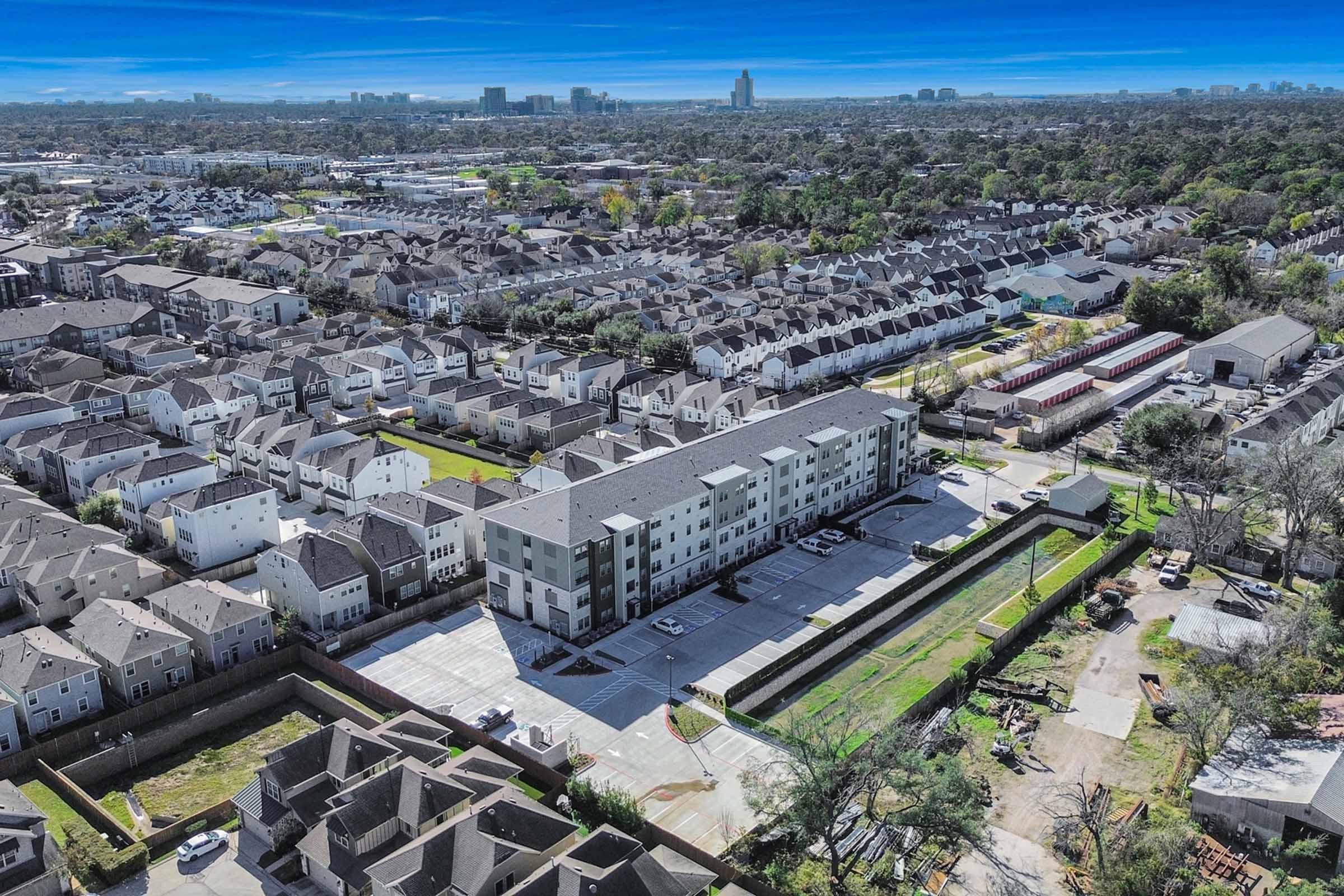 Aerial view of a suburban neighborhood featuring a mix of residential buildings, including multi-family apartments and townhouses. The area is densely populated with greenery and cityscape visible in the background, showcasing a blend of urban and residential environments.