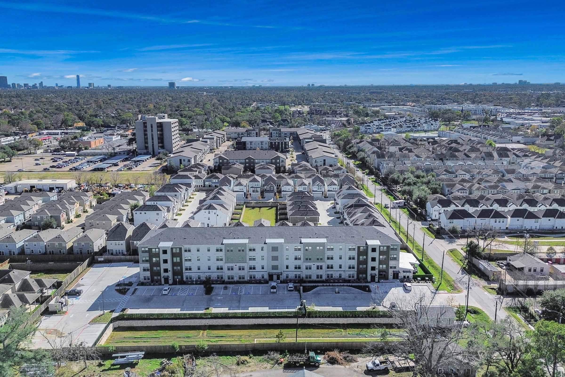 Aerial view of a suburban area featuring multiple residential buildings, including a modern apartment complex in the foreground, surrounded by rows of houses and green spaces. In the background, a city skyline is visible against a clear blue sky.