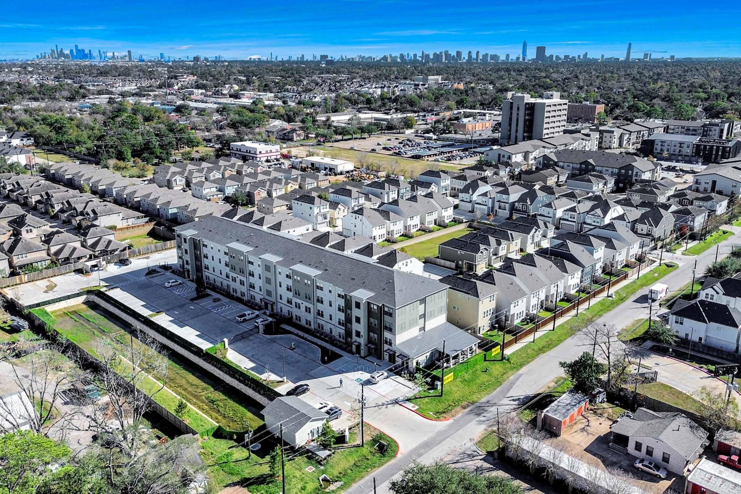 Aerial view of a residential area featuring several multi-story apartment buildings and single-family homes. In the background, a skyline of a city is visible under a clear blue sky, showcasing urban development amidst green spaces.