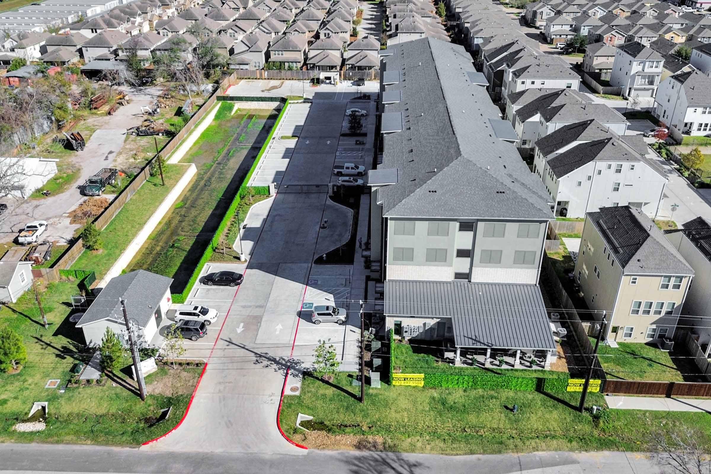 Aerial view of a residential area featuring a multi-unit apartment building with a parking lot beside it. Surrounding the building are well-maintained lawns and a row of houses in the background. The parking area has several parked cars, and the layout reflects a planned community environment.