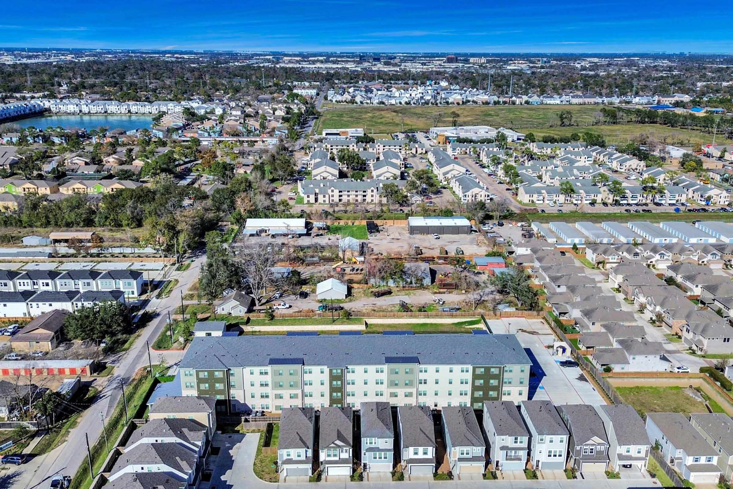 Aerial view of a suburban landscape featuring a mix of residential buildings, green spaces, and roads. Several two- to three-story apartment complexes are visible, along with single-family homes, trees, and patches of farmland in the background, with clear blue skies overhead.