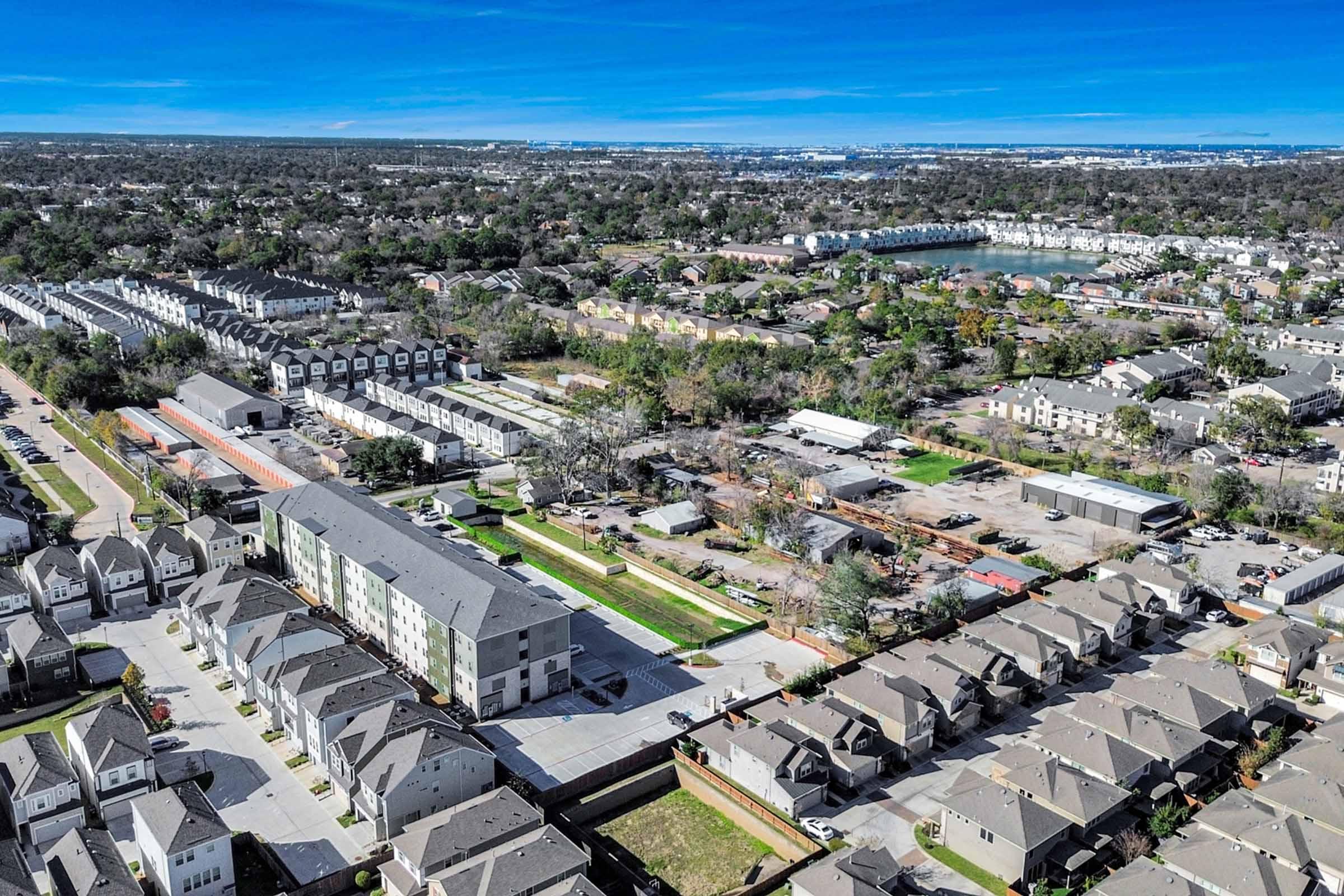 Aerial view of a suburban area featuring several residential buildings and townhouses. The scene includes green spaces, roads, and a nearby lake. The landscape is dotted with trees and various types of housing, showcasing a blend of urban and natural environments. Clear blue skies add to the picturesque view.