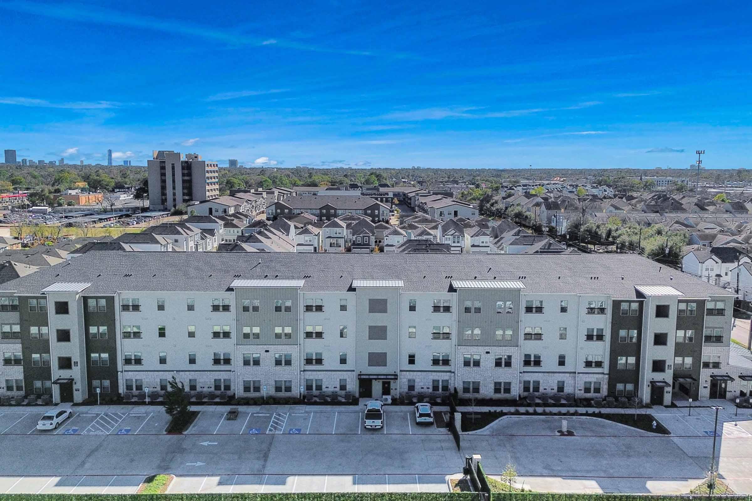 Aerial view of a modern multi-story residential building with a light-colored façade, surrounded by other similar structures and green trees. The scene includes an open parking lot in front, blue sky, and distant cityscape in the background.