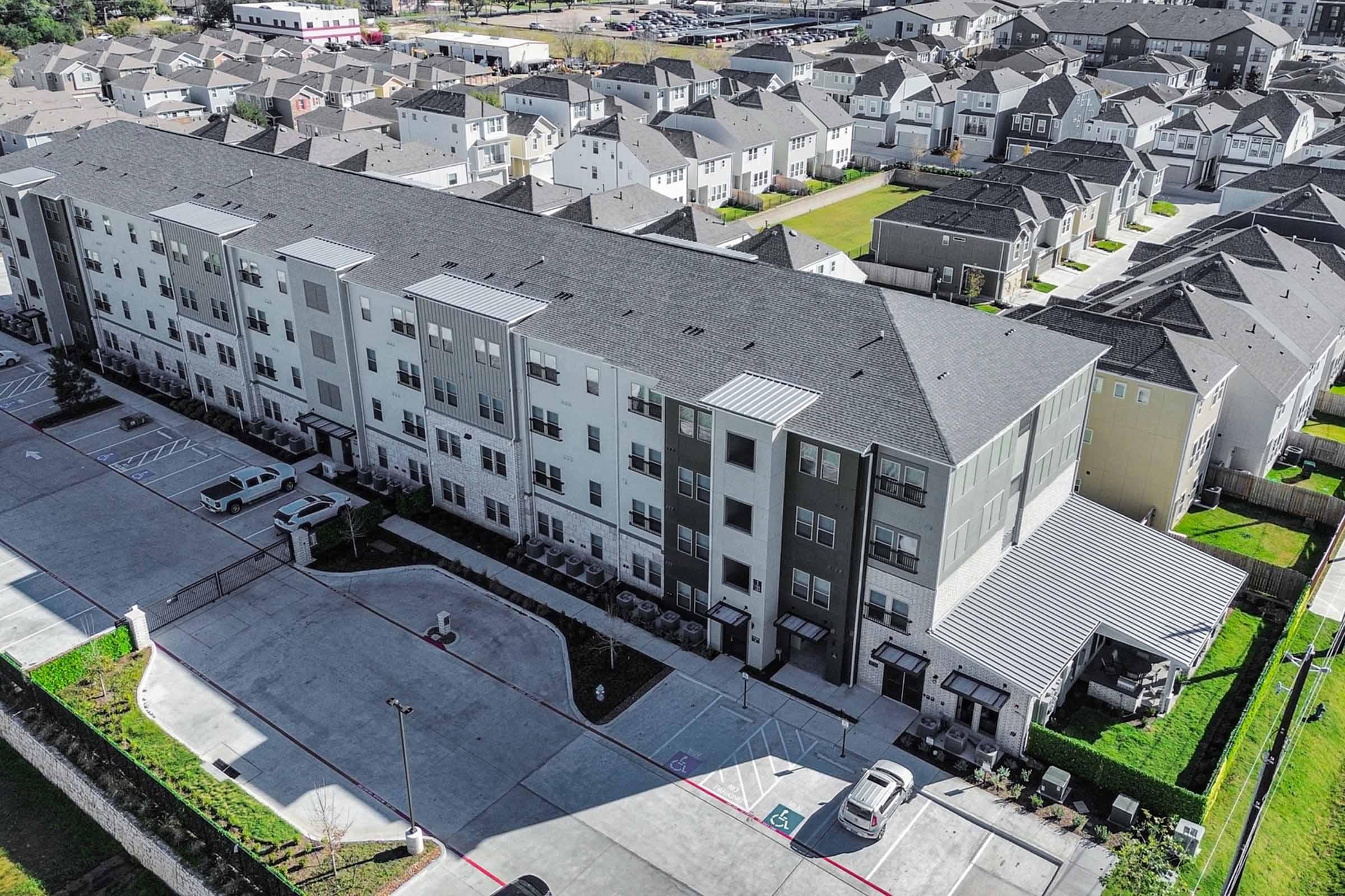 Aerial view of a multi-story residential building surrounded by smaller houses. The building features a mix of grey and beige exteriors, with a parking area in the front. The surrounding area includes more housing, green lawns, and driveways, indicating a suburban neighborhood.