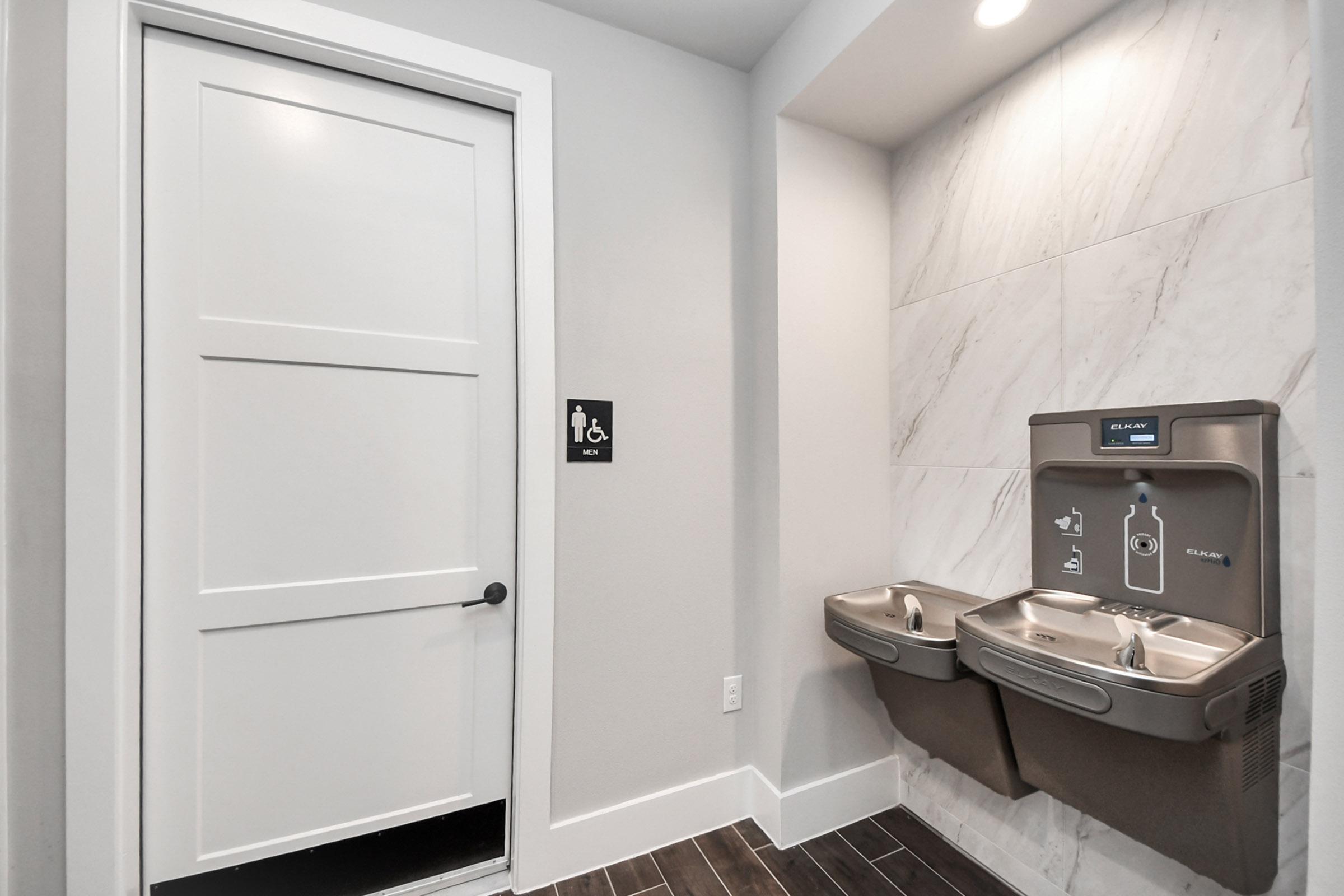 Interior view of a public restroom featuring a closed door with a gender-neutral sign. To the right, there are two stainless steel water fountains mounted on a tiled wall. The area has light-colored walls and dark wooden flooring.