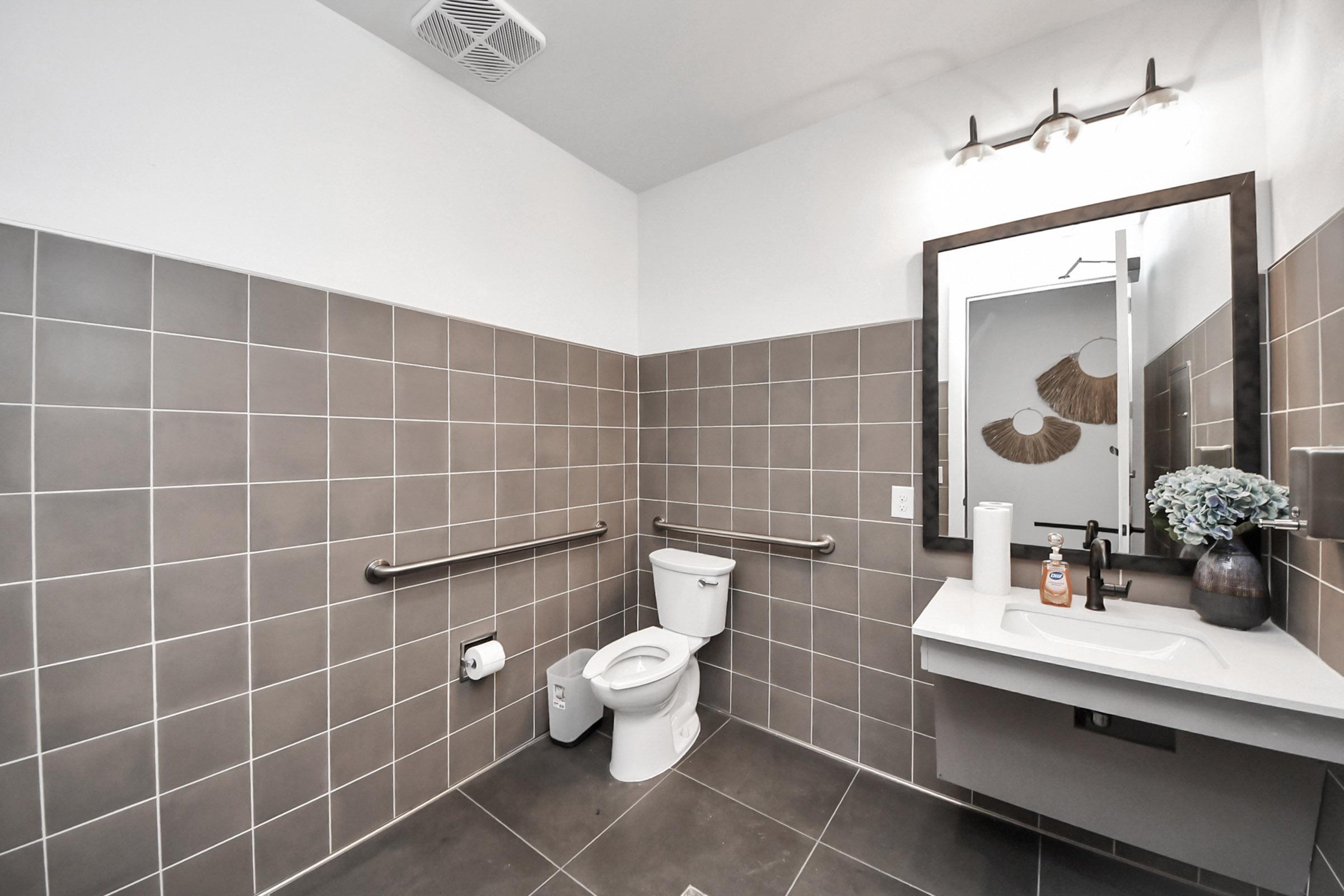 Modern bathroom with large gray tiled walls and dark flooring. Features a white toilet, a wall-mounted sink with a soap dispenser, a large mirror, and grab bars for accessibility. Soft lighting is provided by a fixture above the mirror, creating a clean and functional space.