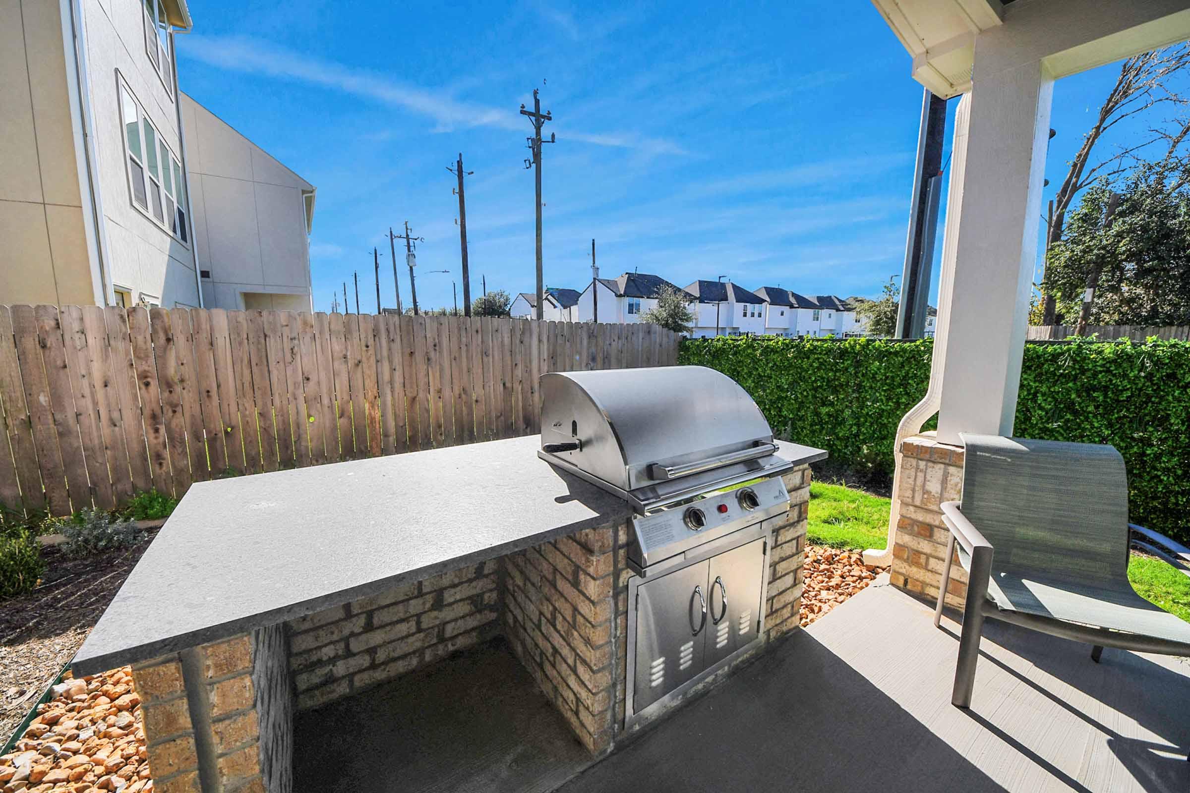 Outdoor grill setup featuring a stainless steel barbecue on a stone surface. The area is surrounded by a wooden fence and lush greenery. In the background, there are residential buildings and utility poles against a clear blue sky. A patio chair is positioned next to the grill for seating.