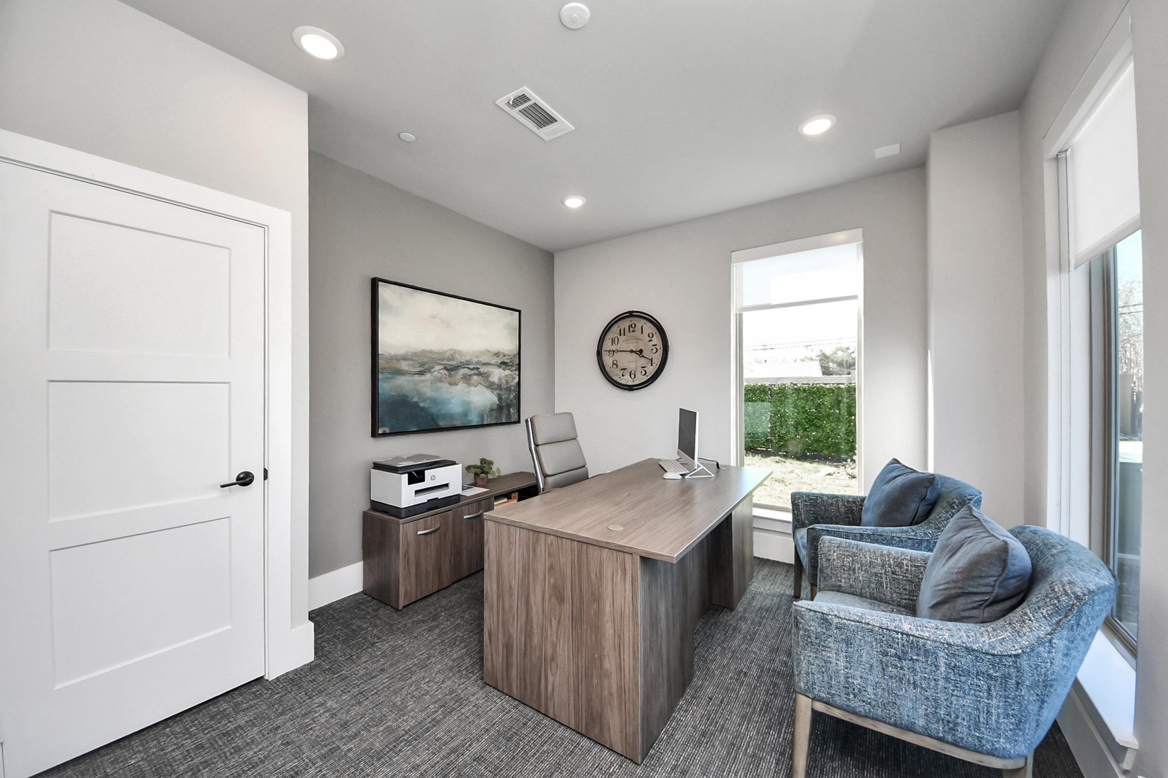 A modern office space featuring a wooden desk with a computer, a printer, two blue armchairs, a large wall clock, and a landscape painting. Natural light enters through a window, while green foliage is visible outside. The walls are painted in a light color, creating a bright and inviting atmosphere.