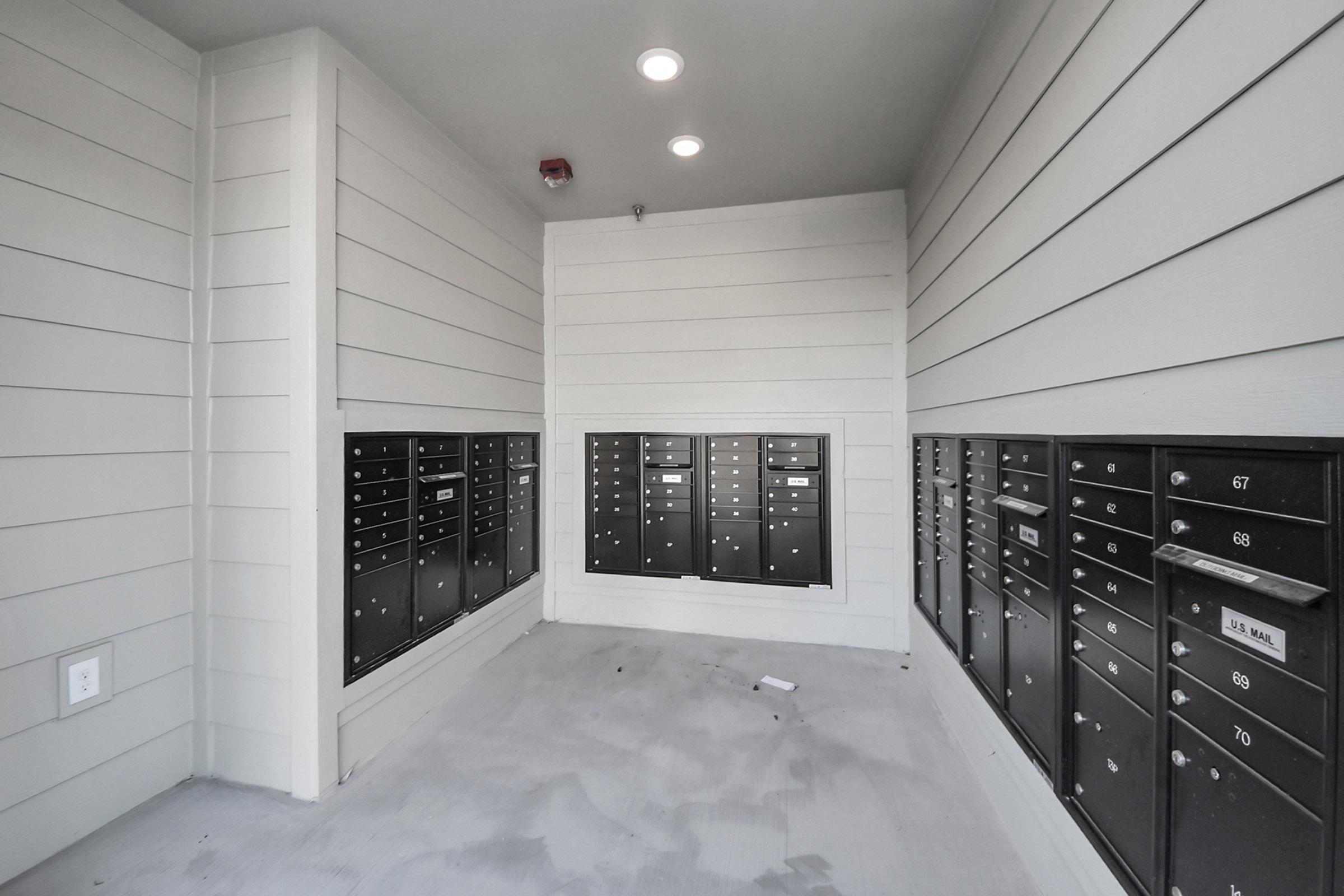 Interior view of a modern mailroom featuring multiple rows of black mailboxes mounted on light-colored walls. The space is well-lit with overhead lights, and the floor is bare with minimal decor.