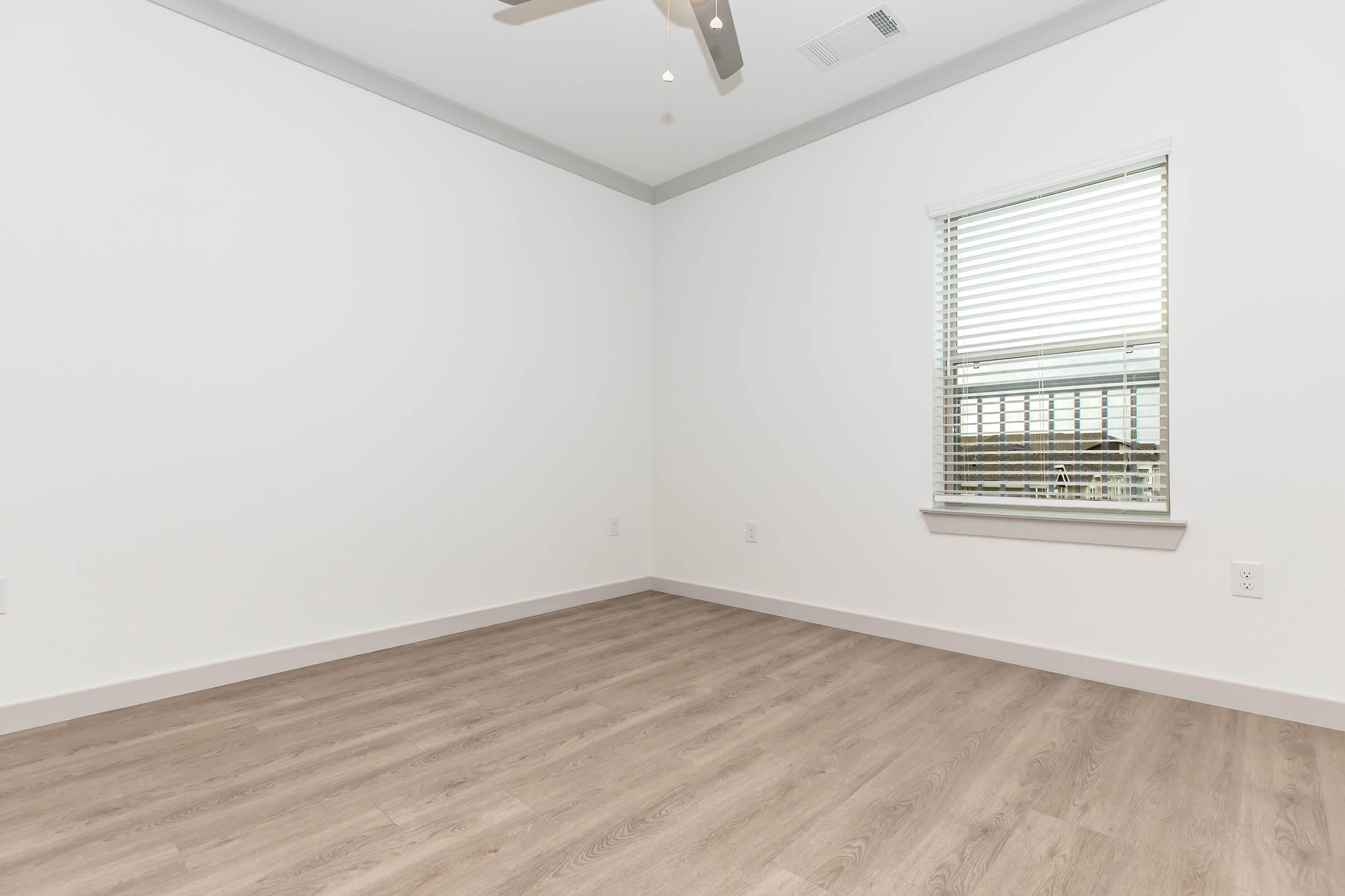 Interior view of a minimalist room featuring light-colored walls, a window with blinds, and wood-like flooring. A ceiling fan is visible, and the space is empty, giving it a clean and open feel. Natural light enters through the window, illuminating the room.