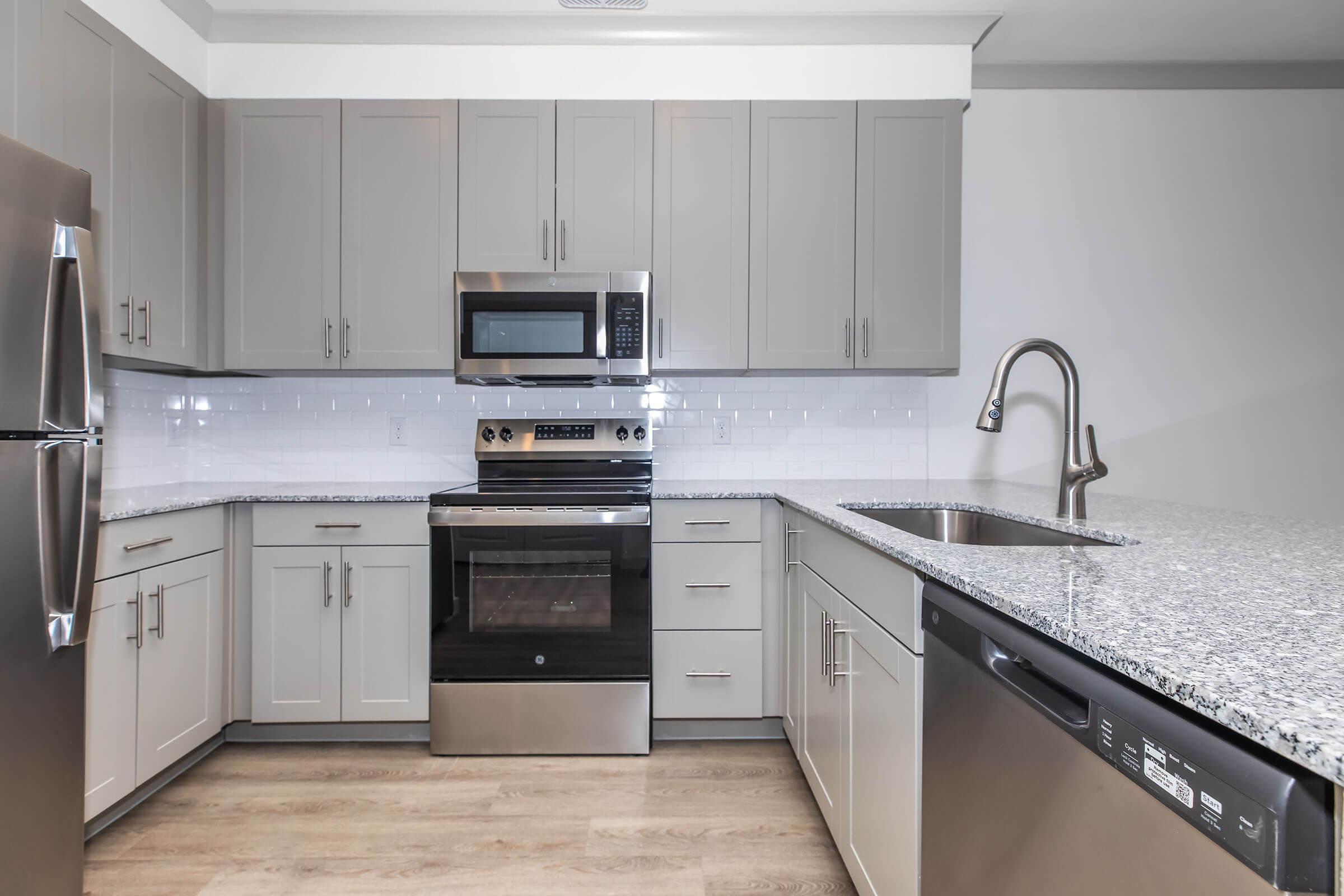 Modern kitchen featuring gray cabinets, stainless steel appliances, a granite countertop, and a sleek faucet. The room includes a microwave and an oven, with a dishwasher located beneath the counter. The flooring is light-colored, adding to the contemporary feel of the space.