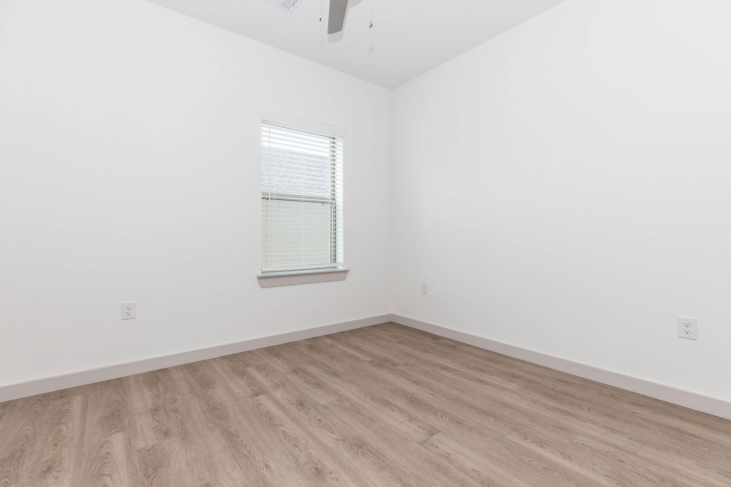 A bright, empty room with light-colored walls and a window. The floor features light wood laminate, and there is a ceiling fan. The space is minimalist, offering a clean and airy atmosphere.