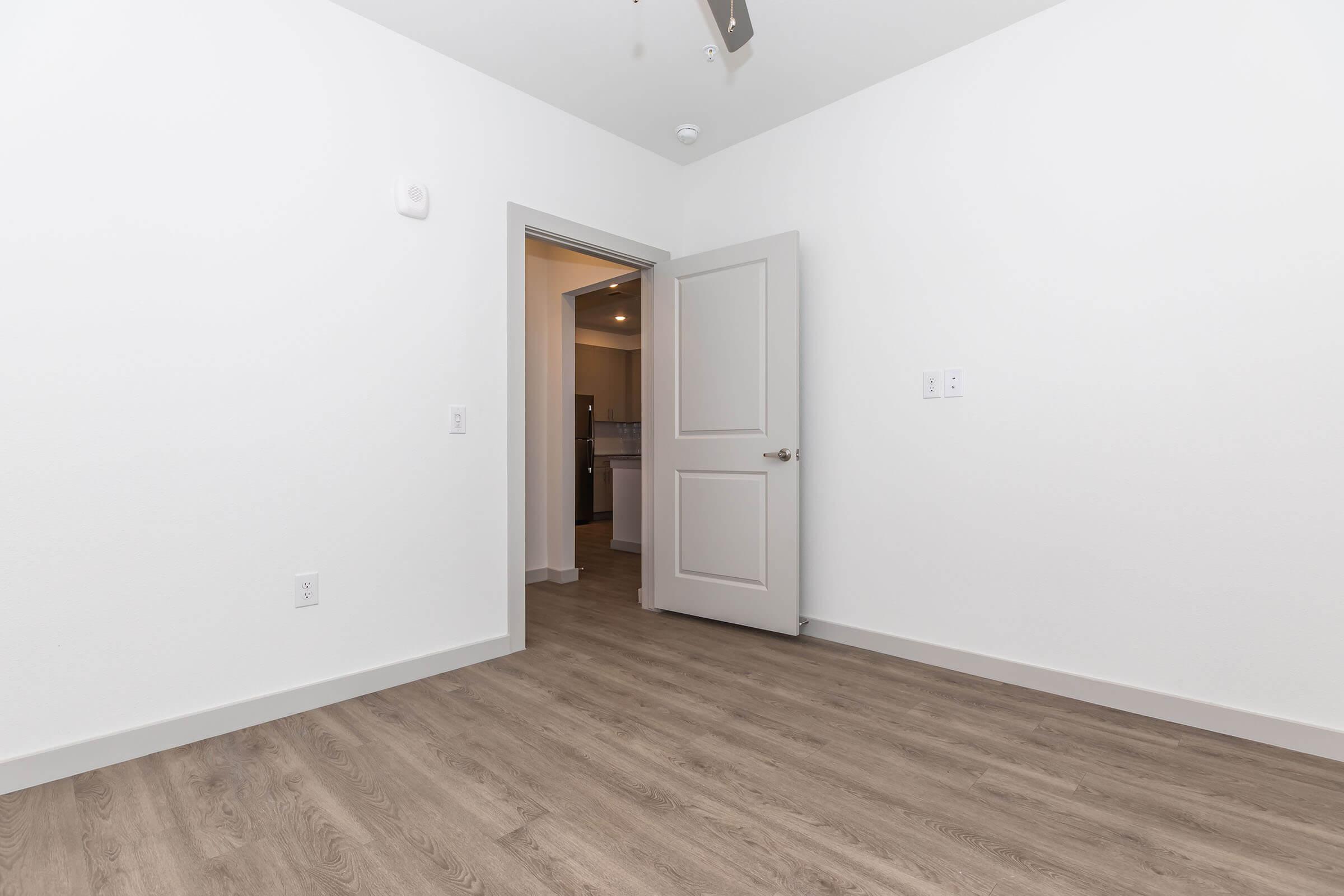 A well-lit, empty room with white walls and a wooden floor. A partially open door leads to another space, and a ceiling fan is visible. The simplicity of the decor suggests it may be a new or recently renovated living area.