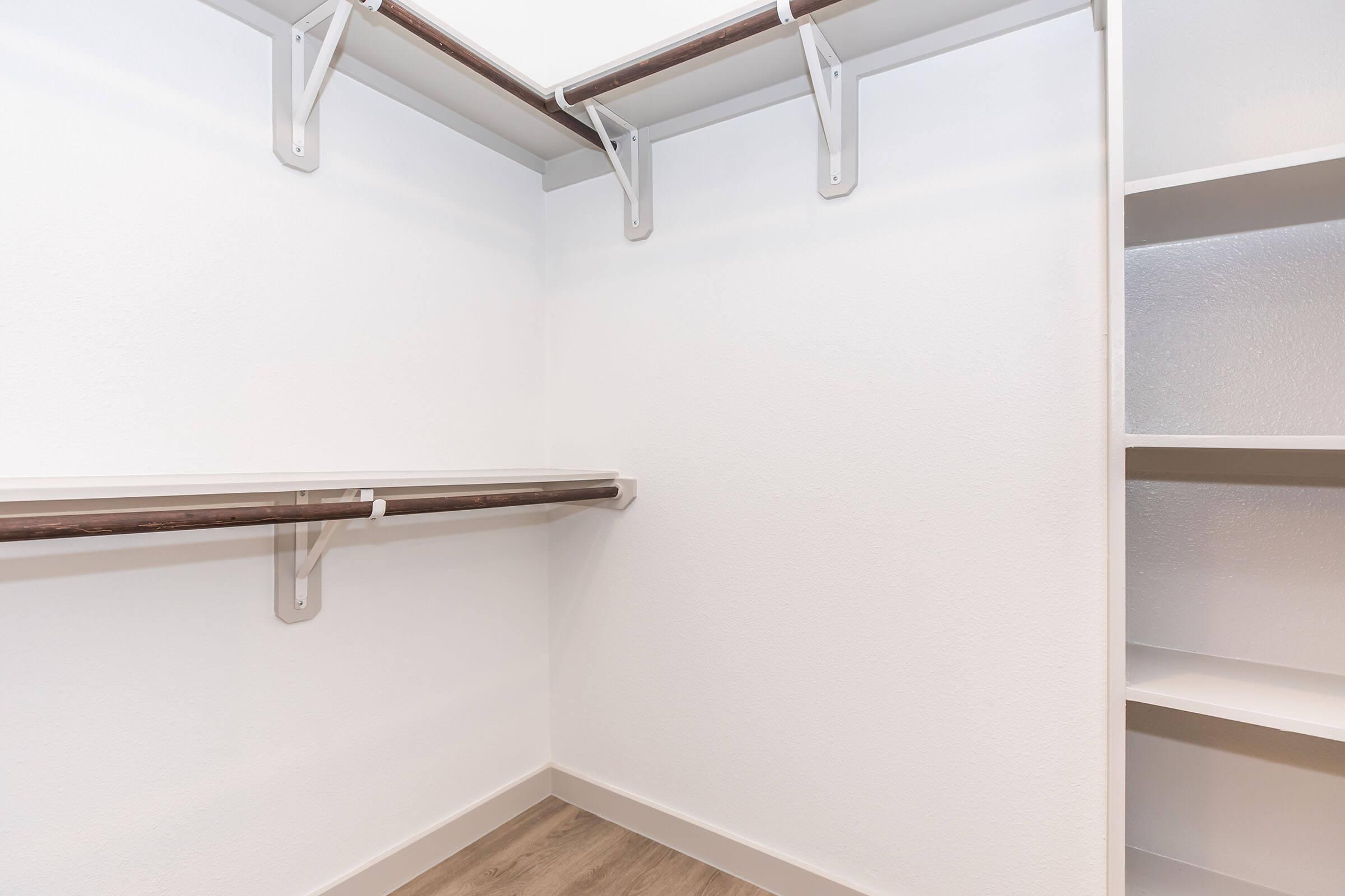 Empty walk-in closet featuring white walls, a light wood floor, and wooden shelves mounted on the walls. There are two brackets on the wall supporting a long shelf, and an additional shelving unit is visible in the corner, creating a spacious and organized storage area.