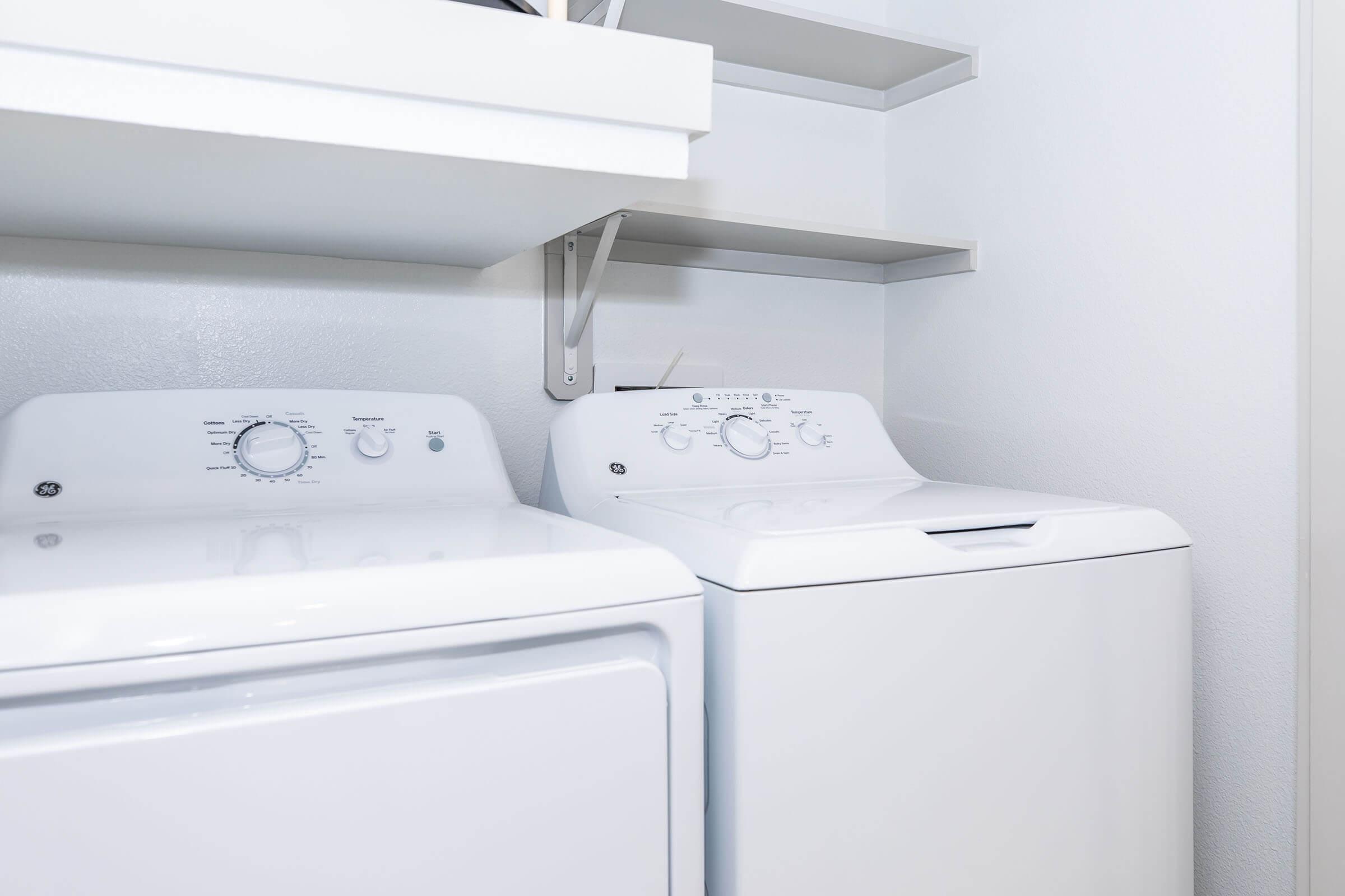 A clean laundry area featuring a white washing machine and dryer side by side, with a simple shelf above them for storage. The walls are white, creating a bright and organized appearance.