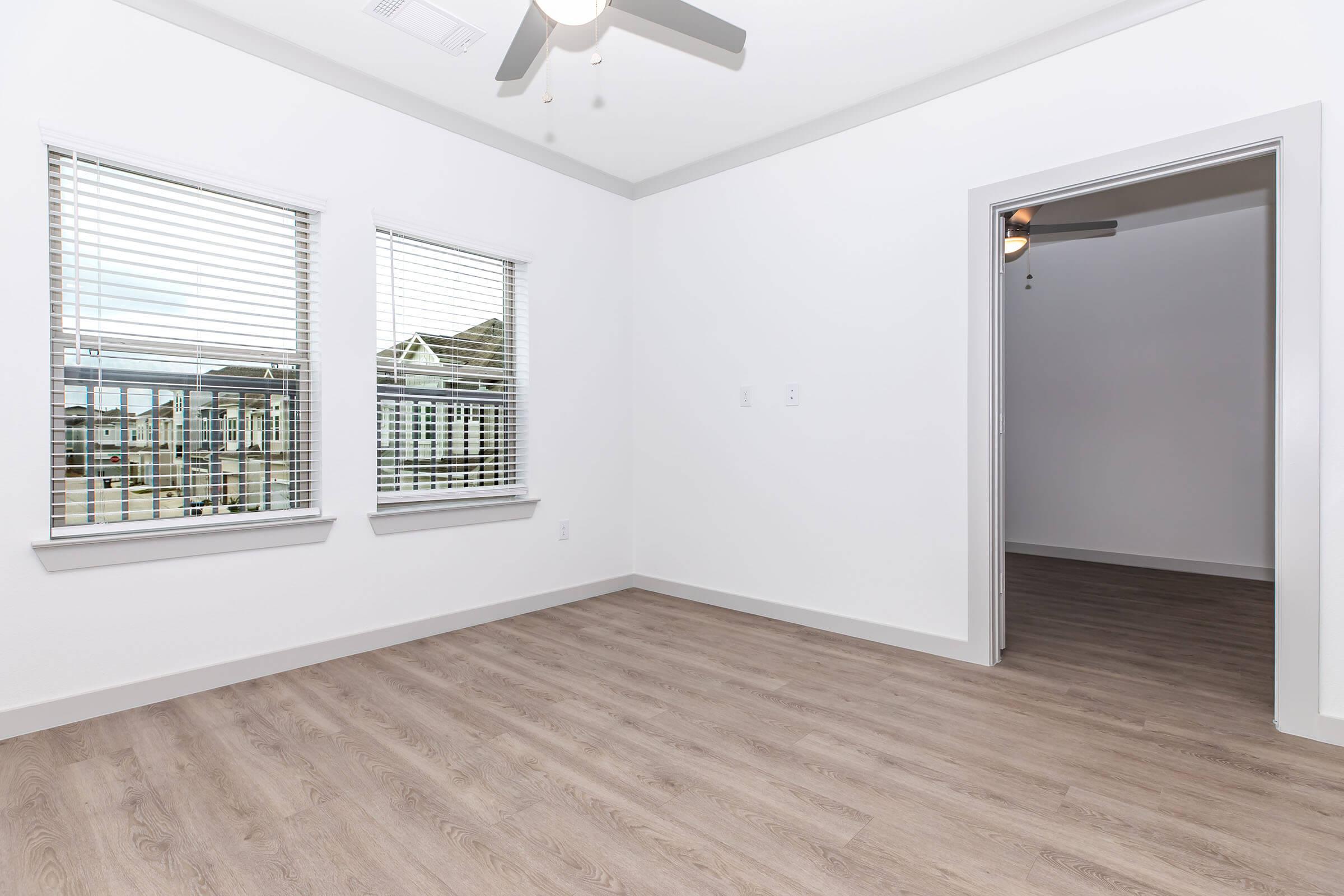 A bright, empty room featuring two windows with blinds, hardwood flooring, and a ceiling fan. The room is painted white, with one wall open to a doorway leading to another space. Natural light illuminates the area, creating a spacious and modern atmosphere.