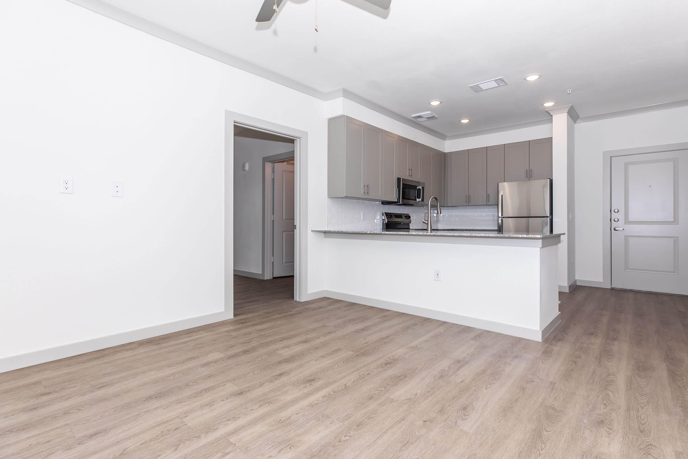 A modern kitchen and living area featuring light wood-like flooring, gray cabinets, stainless steel appliances, and an open layout. A doorway leads to another room, and a ceiling fan is visible. The walls are painted white, enhancing the bright and airy atmosphere of the space.
