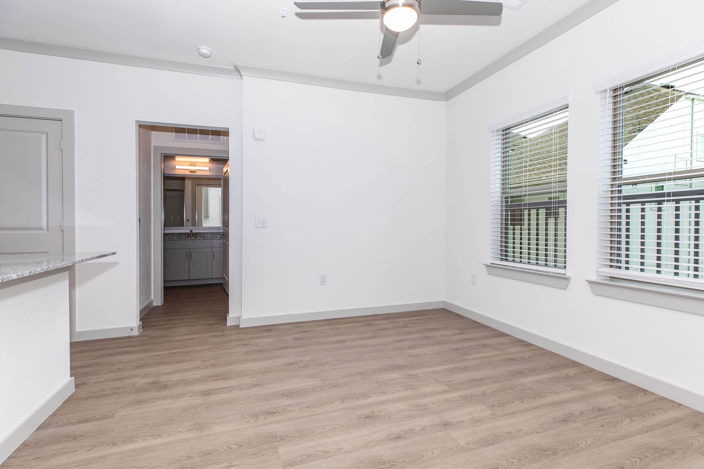 Empty room with light-colored walls and wooden flooring. A ceiling fan hangs from the ceiling. Two windows on the right side let in natural light. In the background, a doorway leads to a bathroom area with a mirror. The overall space appears modern and uncluttered.
