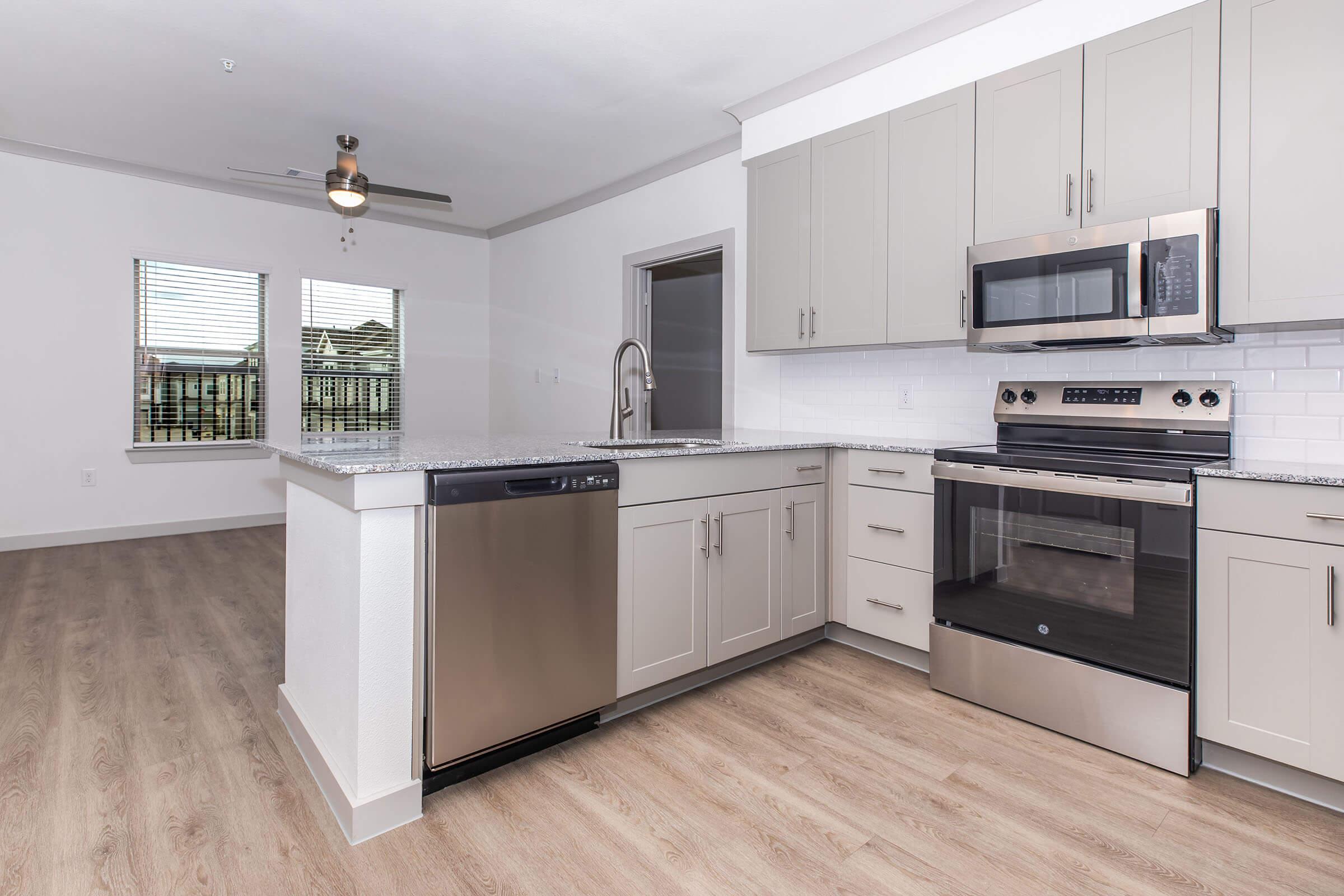 Modern kitchen featuring light gray cabinets, stainless steel appliances including a dishwasher and oven, a granite countertop, and wood-style flooring. Two windows allow natural light, and there is an open layout leading into a living space.