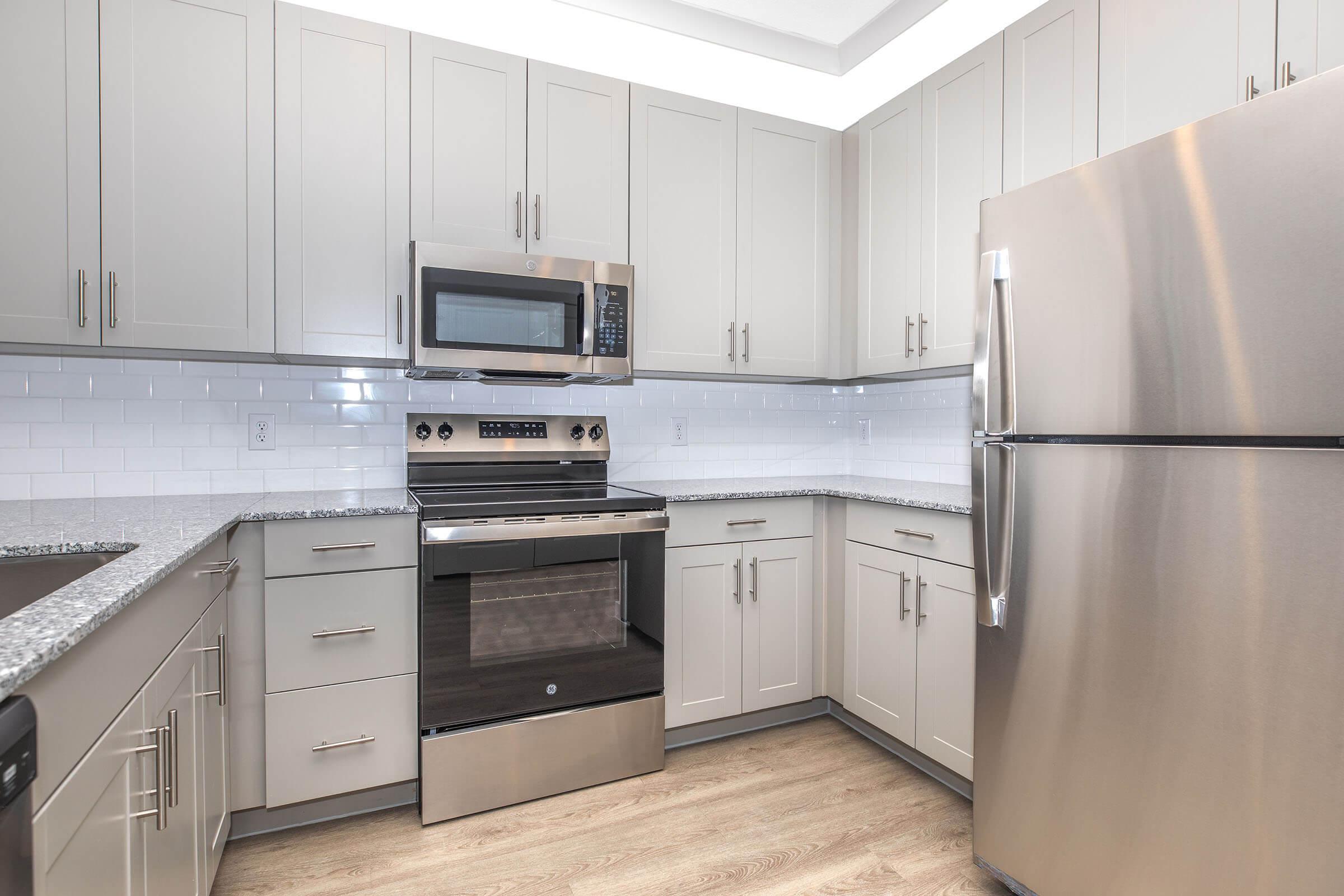 Modern kitchen with light gray cabinets, stainless steel appliances including a microwave, oven, and refrigerator, and a granite countertop. The backsplash features white subway tiles, and the flooring is a light wood finish, creating a sleek and contemporary look.